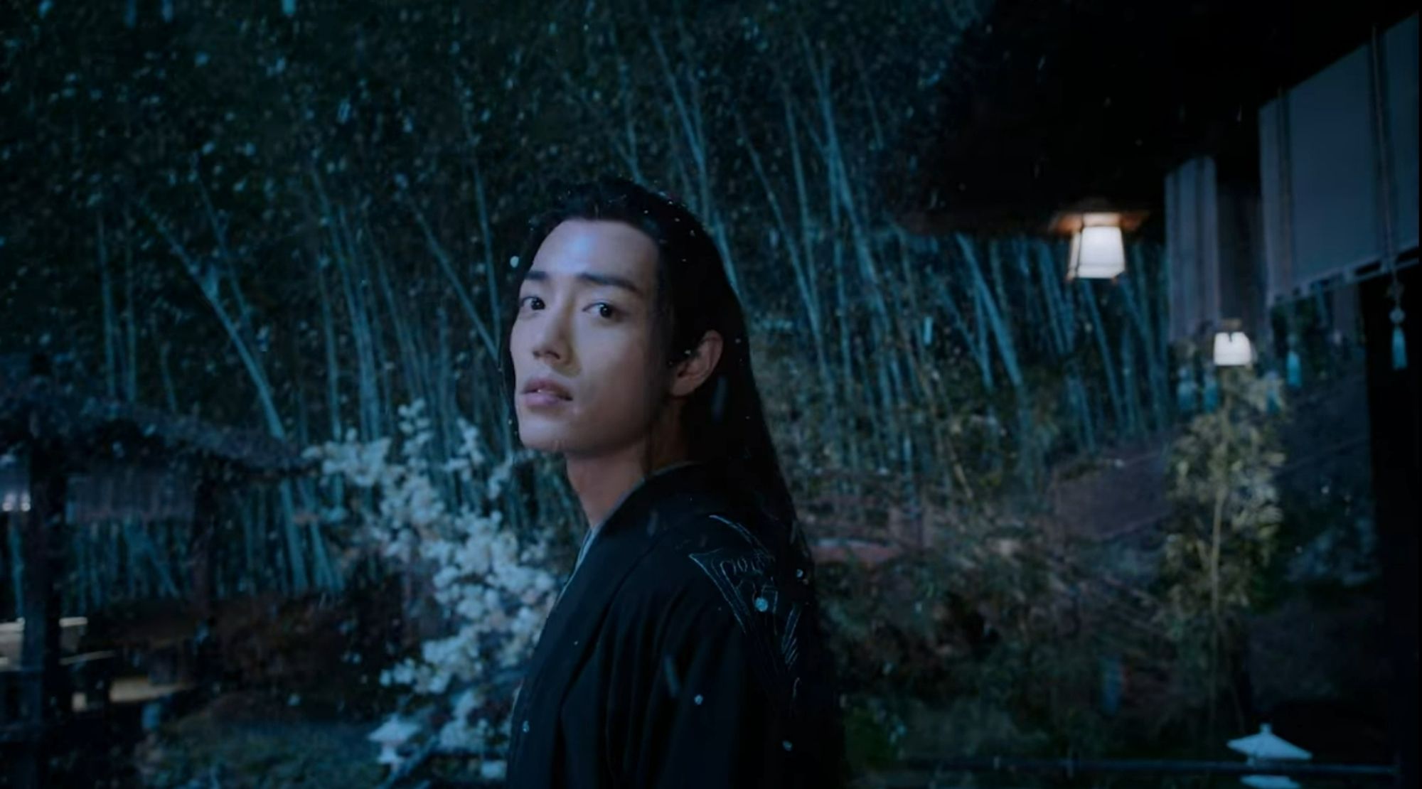 Closeup of Wei Wuxian (actor Xiao Zhan) standing outside while it's snowing, he's turned his head towards the camera. He's in picture from chest up, wearing black robes.