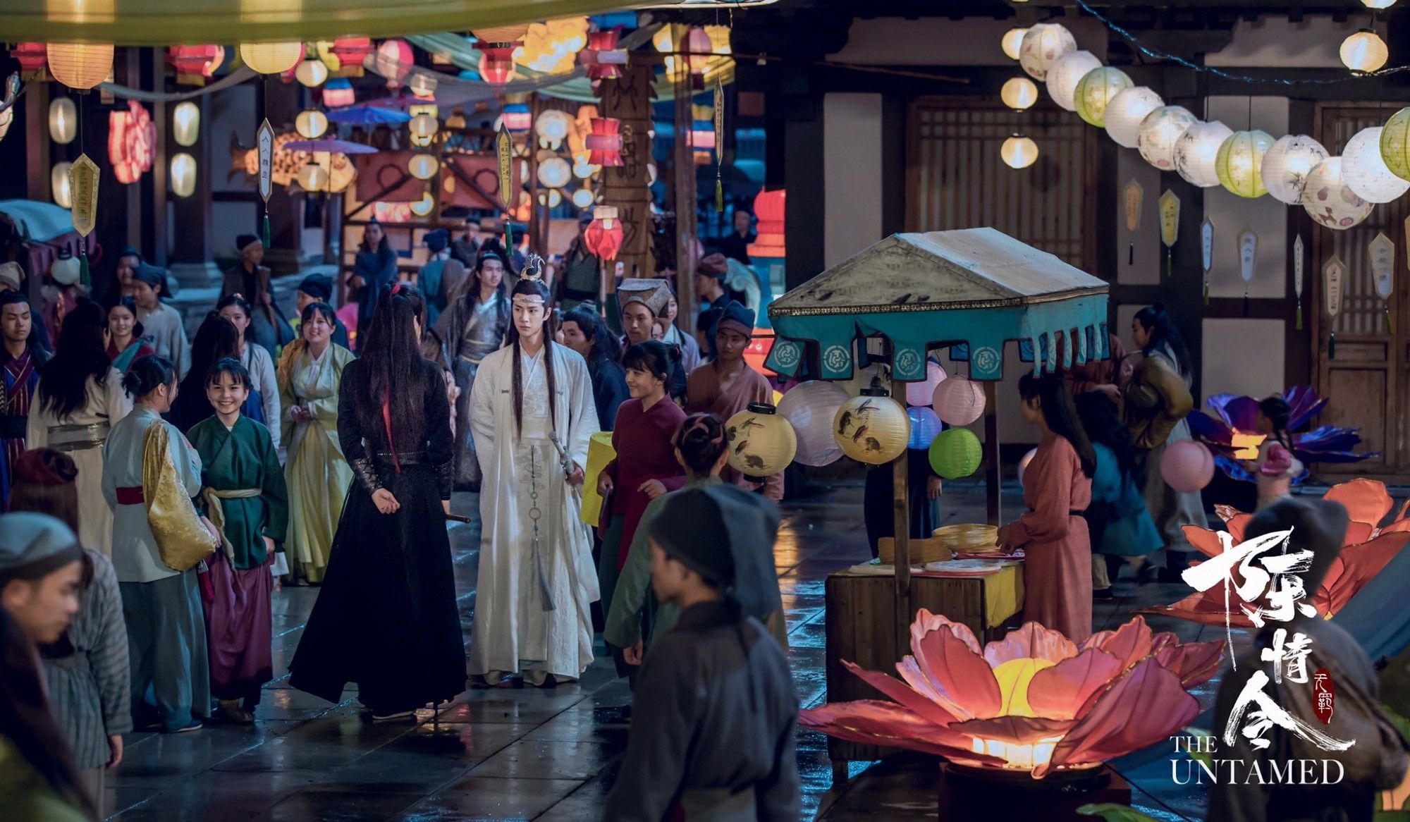 Wei Wuxian (actor Xiao Zhan) and Lan Wangji (actor Wang Yibo) standing in a middle of a town staring at each other. There"s lots of people and colourful lit lantern around. Wei Wuxian in black robes, Lan Wangji in white.