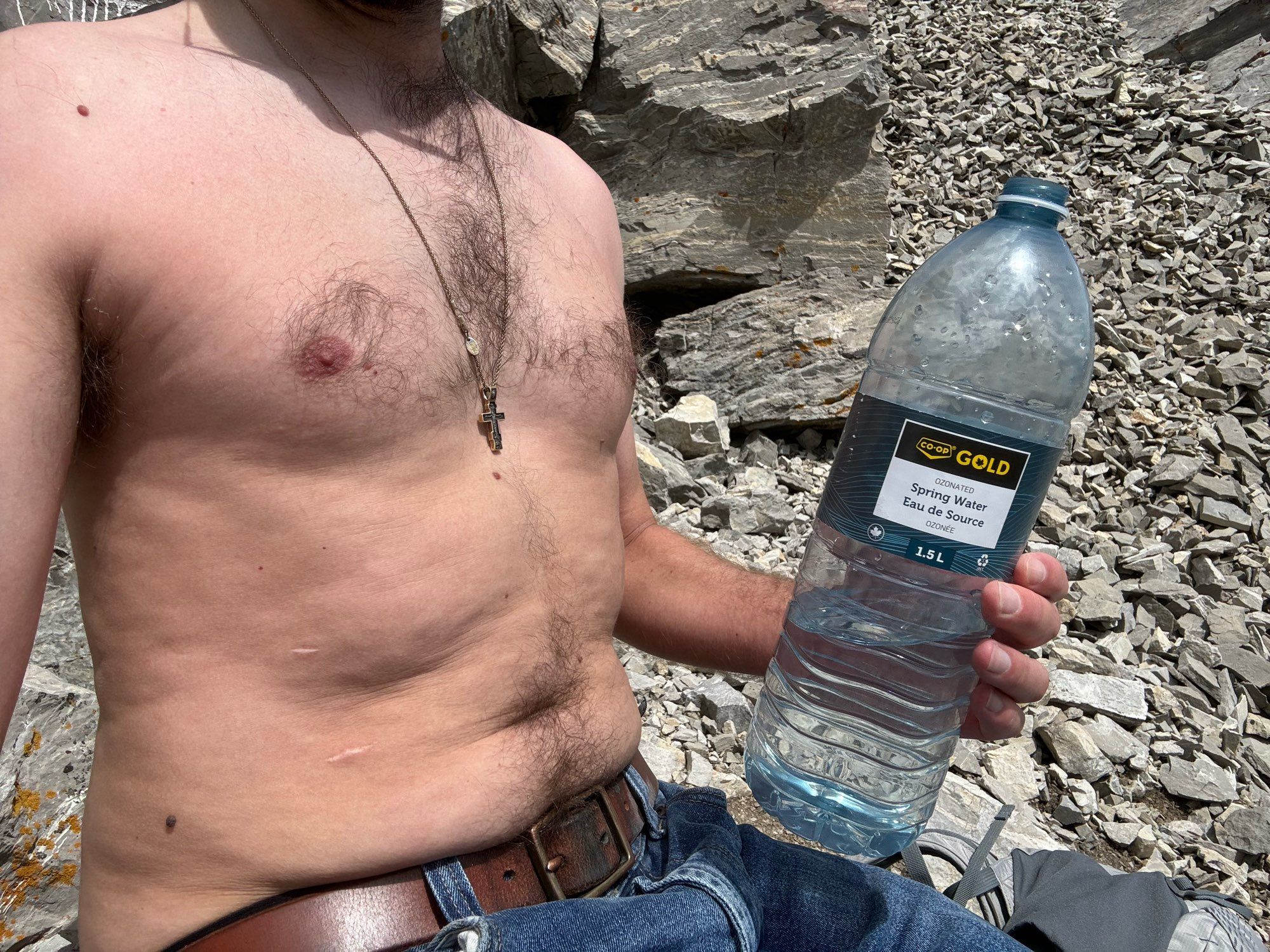 Shirtless male sitting on a rock on a mountain and holding a large bottle of water