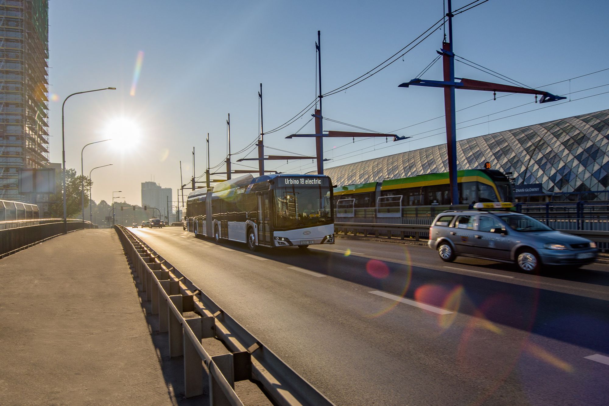 An electric bendy bus on a sunny street with lens flare, modern building and car nearby.

Photo by Solaris