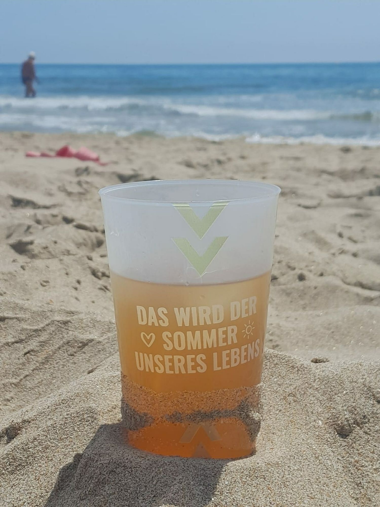 Ein Becher mit der Aufschrift "das wird der Sommer unseres Lebens" steht am Strand im Sand. Er ist gefüllt mit Tequila Sunrise und einem Eiswürfel. Im Hintergrund sieht man das Meer.