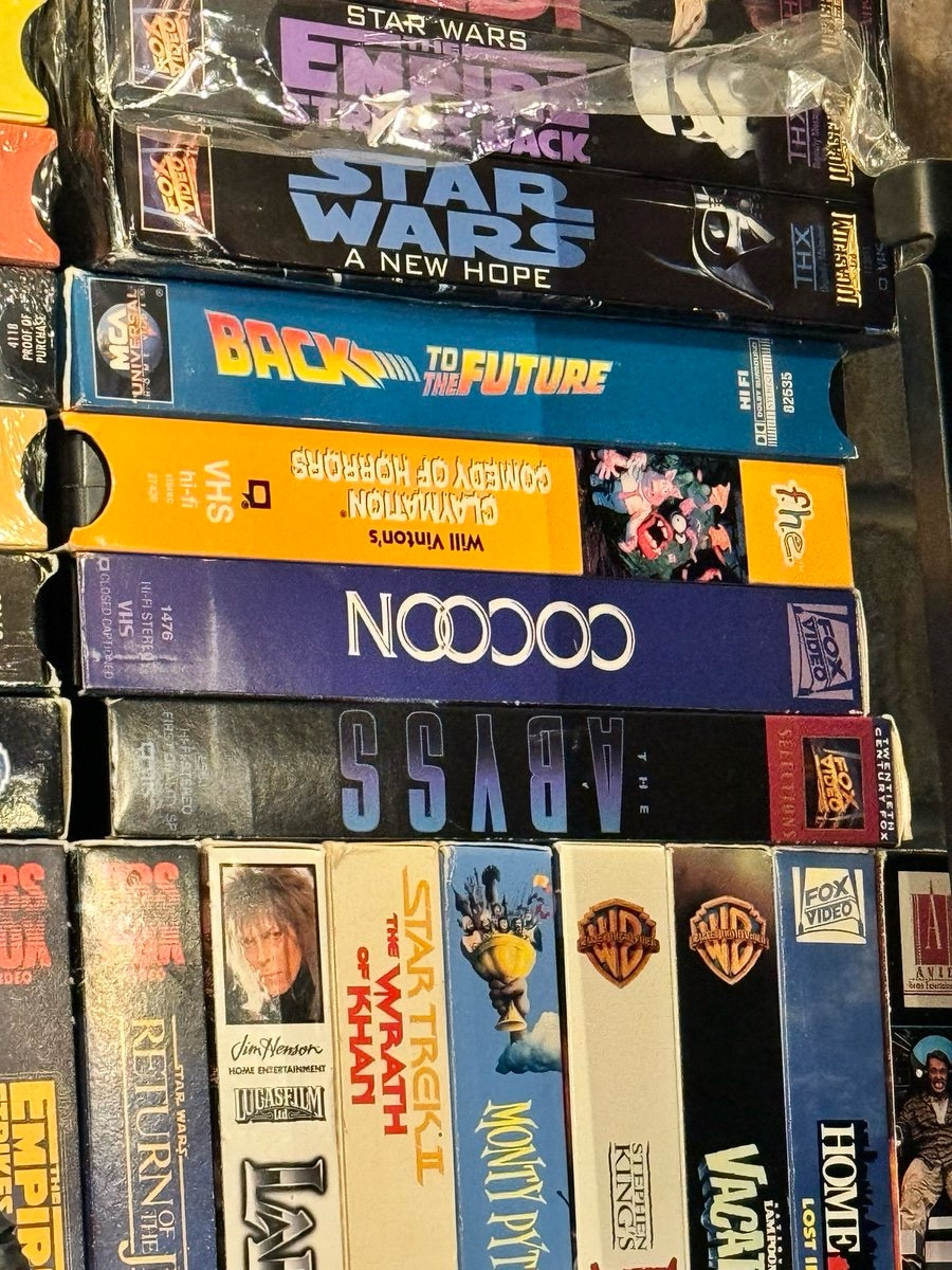 A stack of VHS movies, where the movie Cocoon is set upside down, so it looks like it reads “NOOOOO”