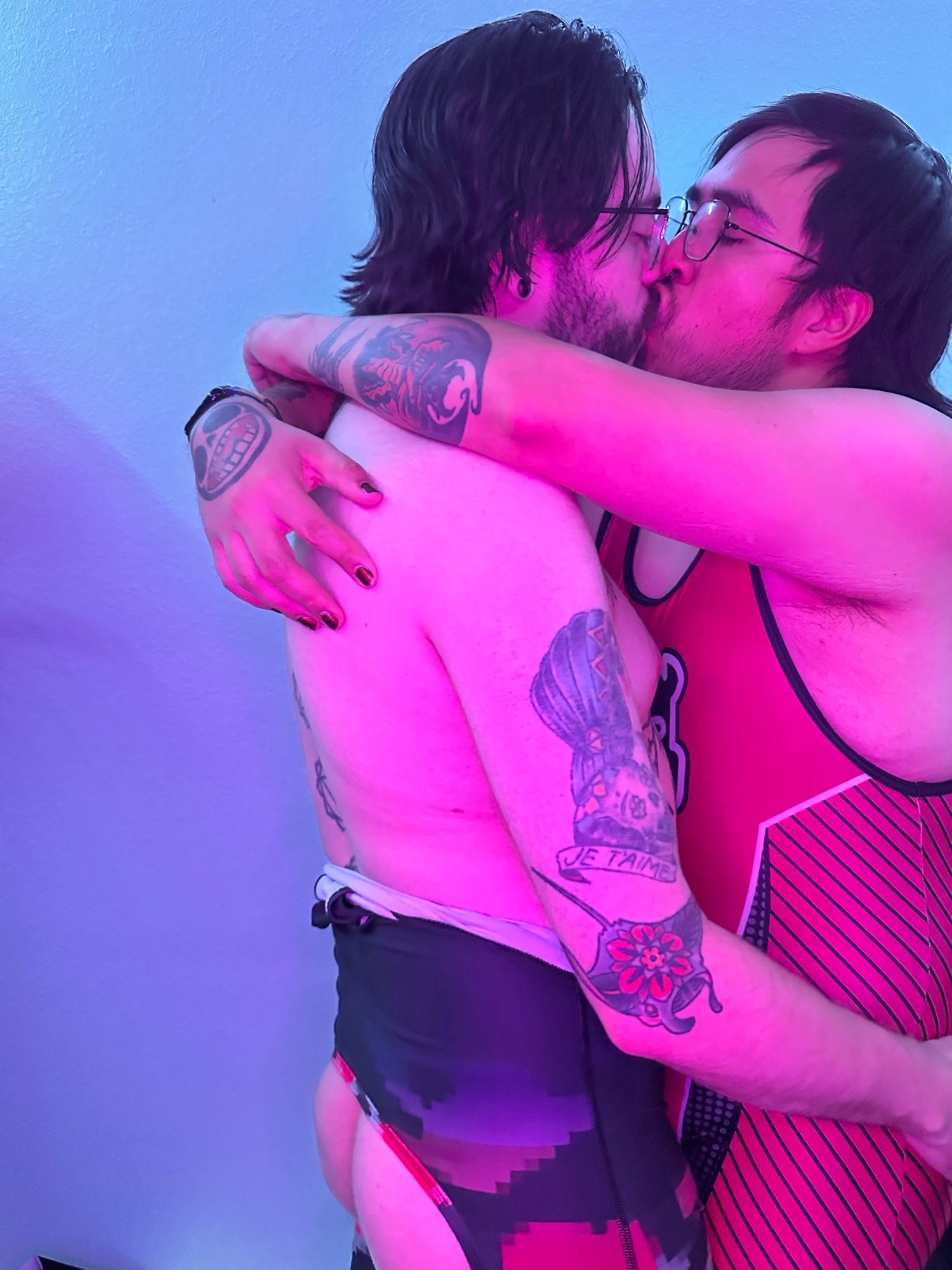 Two people making out. The first on the left in a pink and black singlet pulled down to the waist that is assless and the second in a pink and black stripped singlet