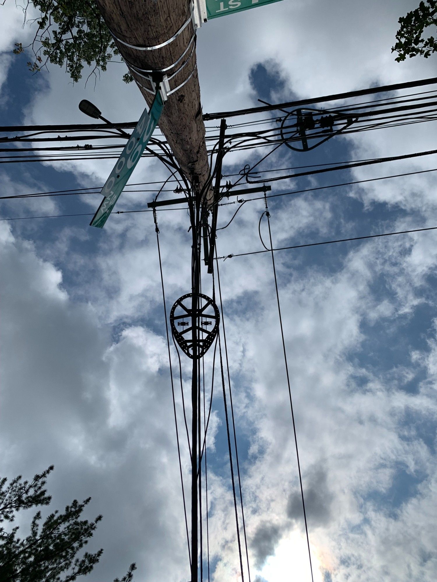 Farther-away view of a telephone and power line array that looks like it could be a mask for a human face. Wires are attached to a pole that contains a street sign. Thin clouds are in the background of the picture, which is taken from the ground looking up.