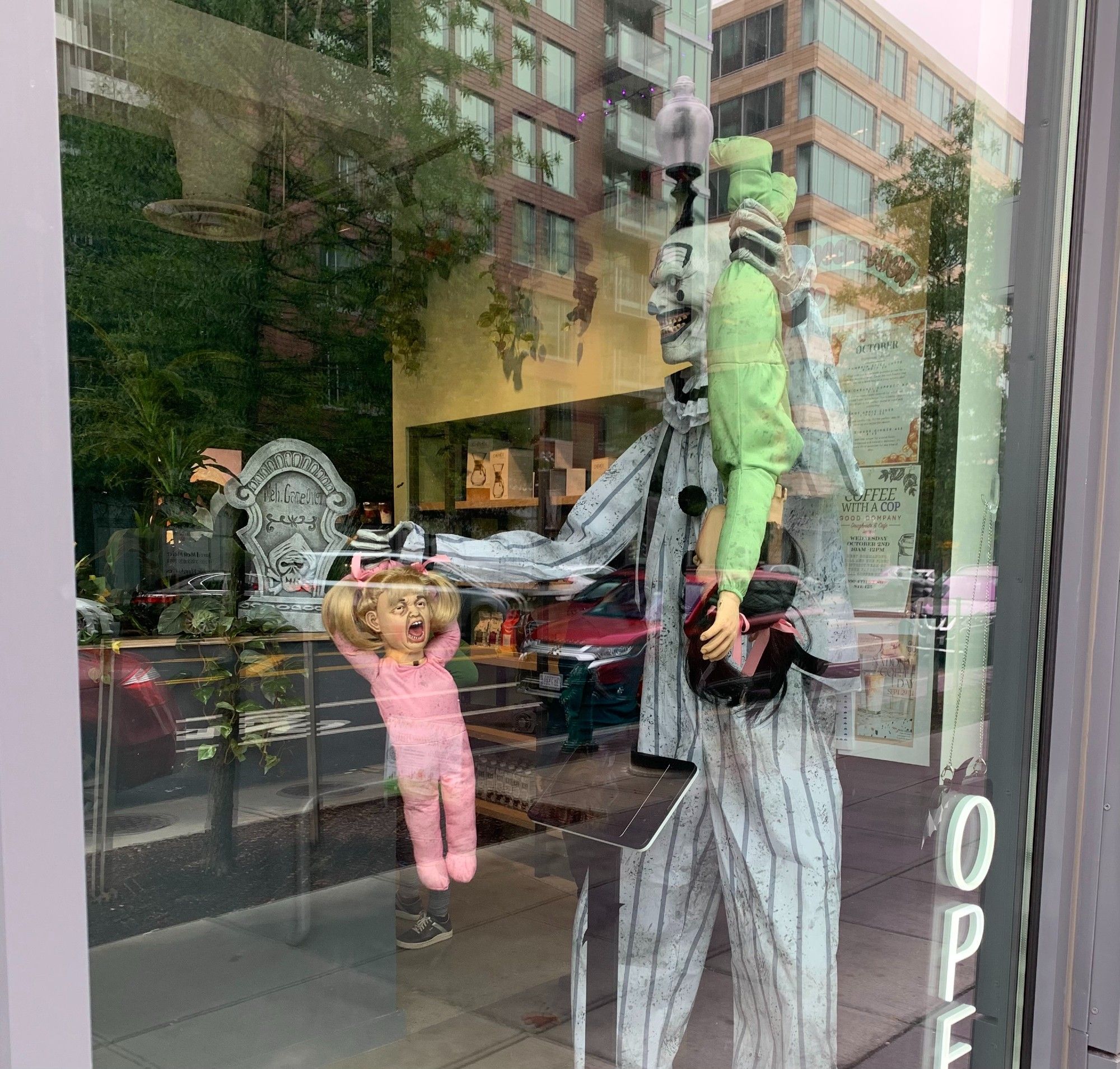 A Halloween decoration in the front window of Good Company Doughnuts & Cafe shows a tall scary clown. The clown holds two toddlers. One is hanging upside down. The other toddler is being held by her hair and appears to be screaming.