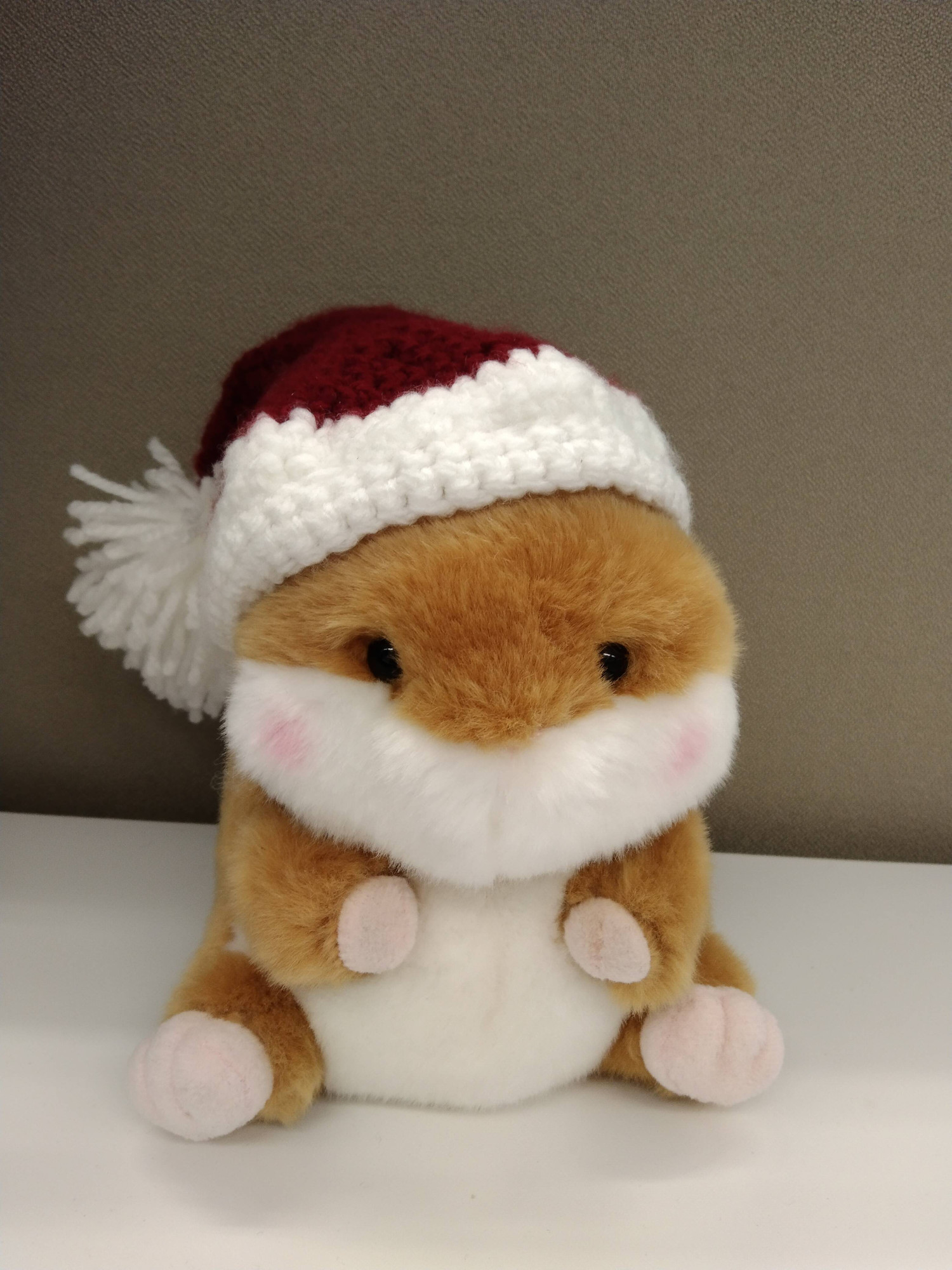 Foto de Bibu, el peluche de hamster suave, de color blanco y naranja y mejillas sonrosadas, con un gorrito de ganchillo puesto. El gorro es parecido al de papá Noel, blanco en los bordes, rojo el resto y con un pompón blanco un peludo en la punta.