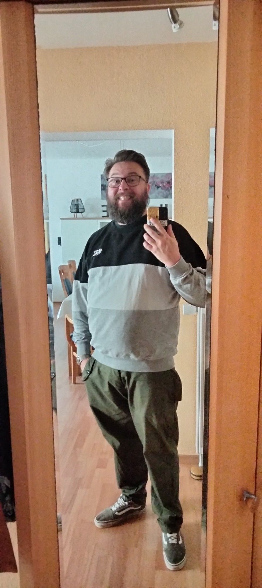 Ich vorm Schrank meiner Eltern, wo ich auf den Autoschlüssel lauere. Ich trage graue Vans, ne olivgrüne Chino und einen von unten nach oben grauweißschwarz dreigestreiften Pullover. Ich halte mein Handy vor meiner rechten Schulter, grinse leicht gezwungen durch meinen flusigen Bart und habe aus Nordseewindgründen eine Föhnfrisur.
Im Hintergrund sieht man einen Miniausschnitt der Einrichtung meiner Eltern. Viel Weiß und Buche erfüllt den Raum, der dadurch sehr hell wirkt.