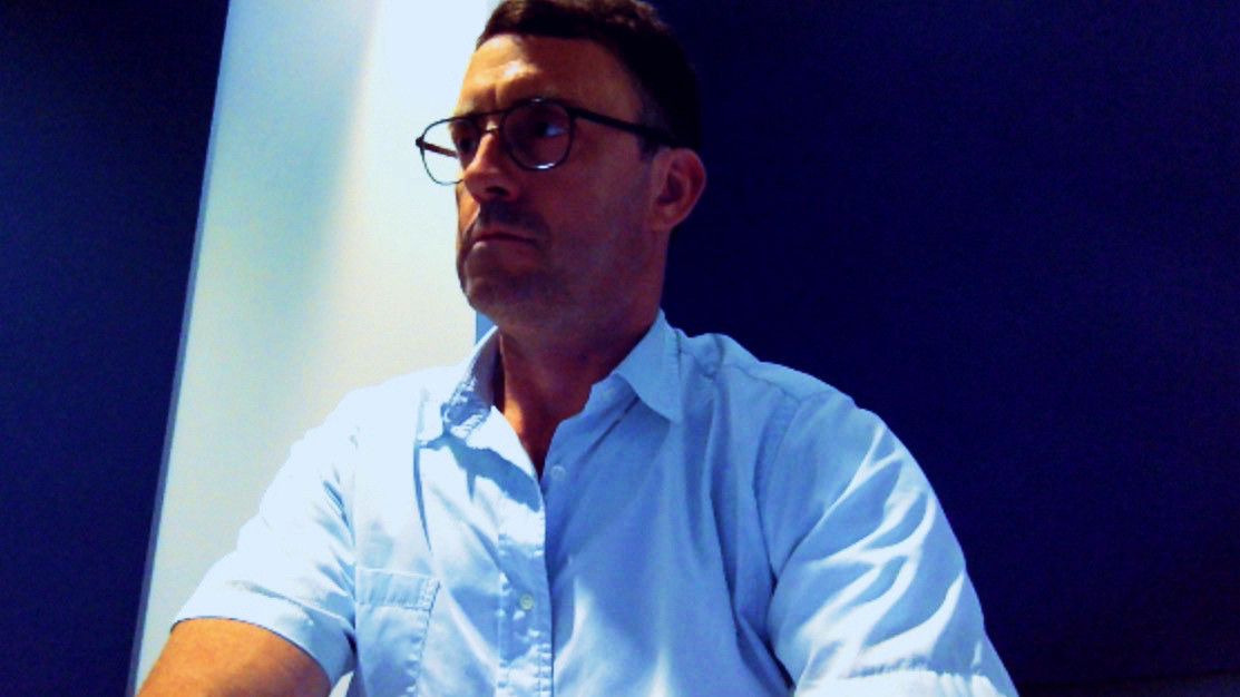 Photo of a man wearing moscot glasses and a blue short sleeve shirt. It has a 1960s NASA control booth vibe, like a film still from a movie. Portrait of Alexander Eiserloh, a man in his 40s (at the time).