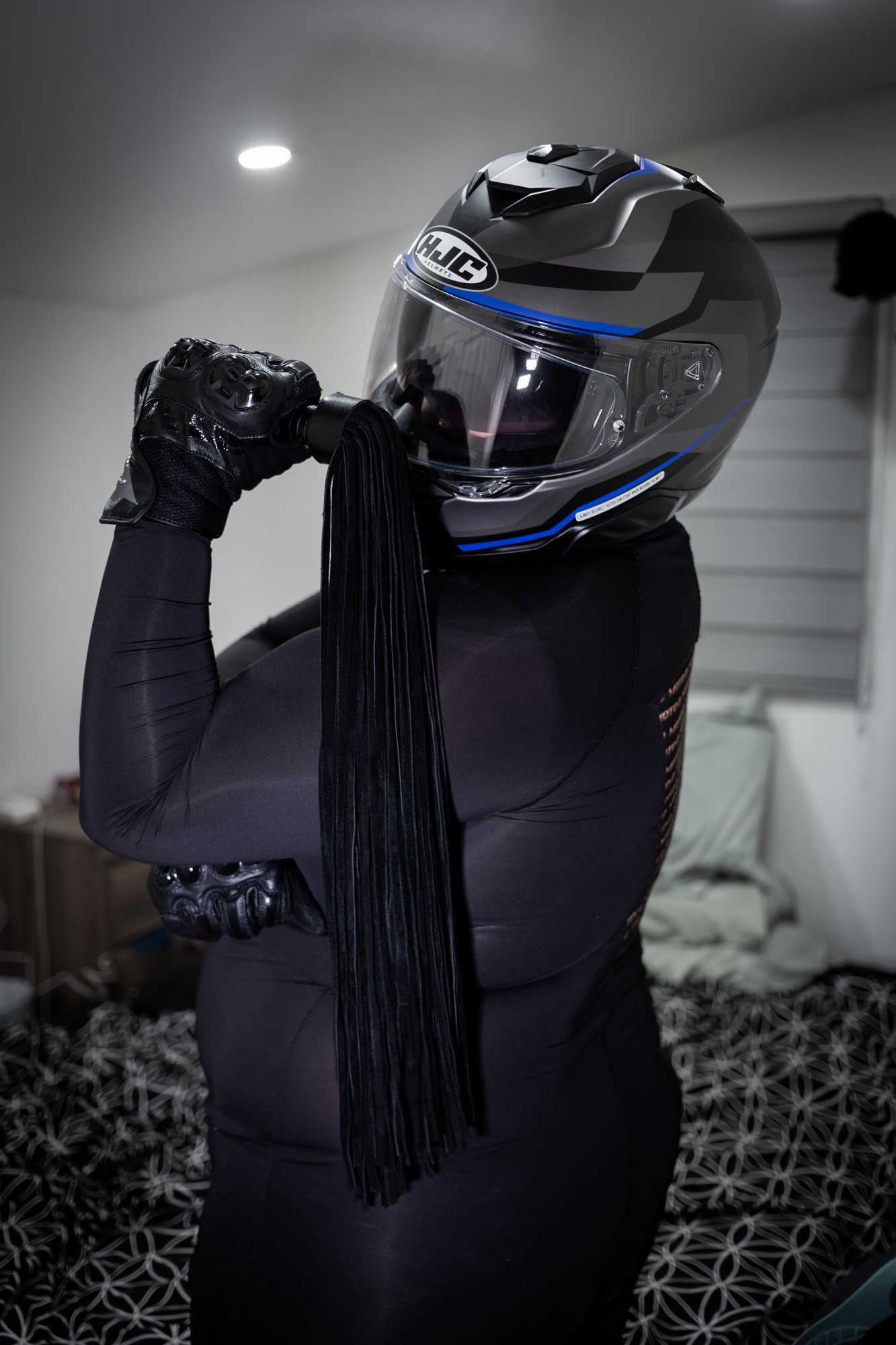 Thigh up photo of me standing in a profile angle. I am wearing a black spandex suit, leather gloves and a full face motorcycle helmet. I am holding a suede flogger over my shoulder.