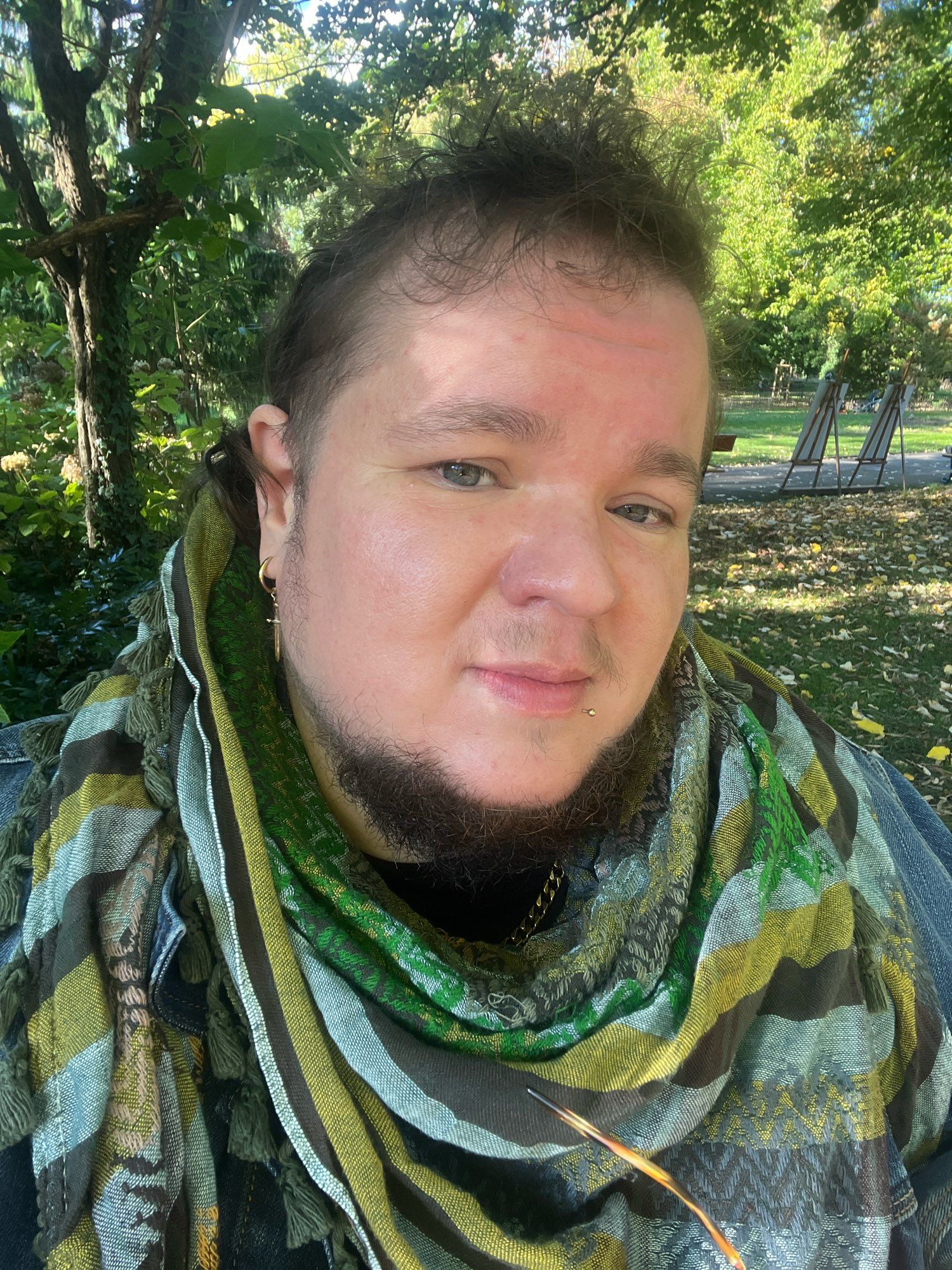 Selfie de moi homme blanc aux yeux bleus, barbe au niveau du menton, coupe mulet, anneau à l’oreille avec charms en forme d’épée, et keffieh palestinien vert, marron et jaune. Je suis dans un parc.