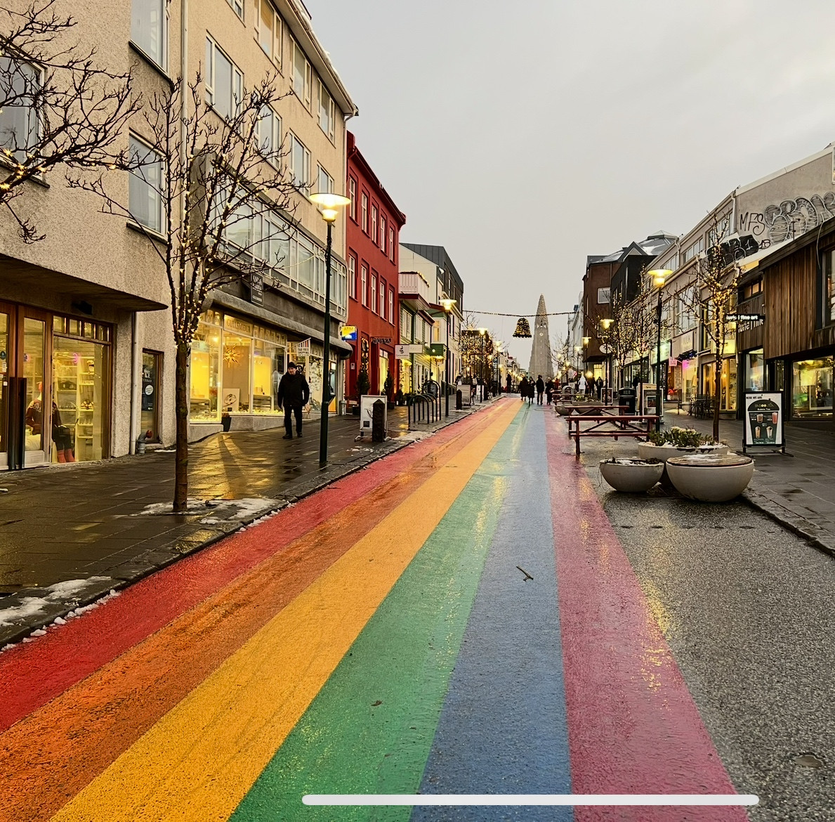 Shopping street in Reykjavik, Iceland in which the drivable portion is painted in broad rainbow stripes, receding to a modern cathedral tower in the distance 
