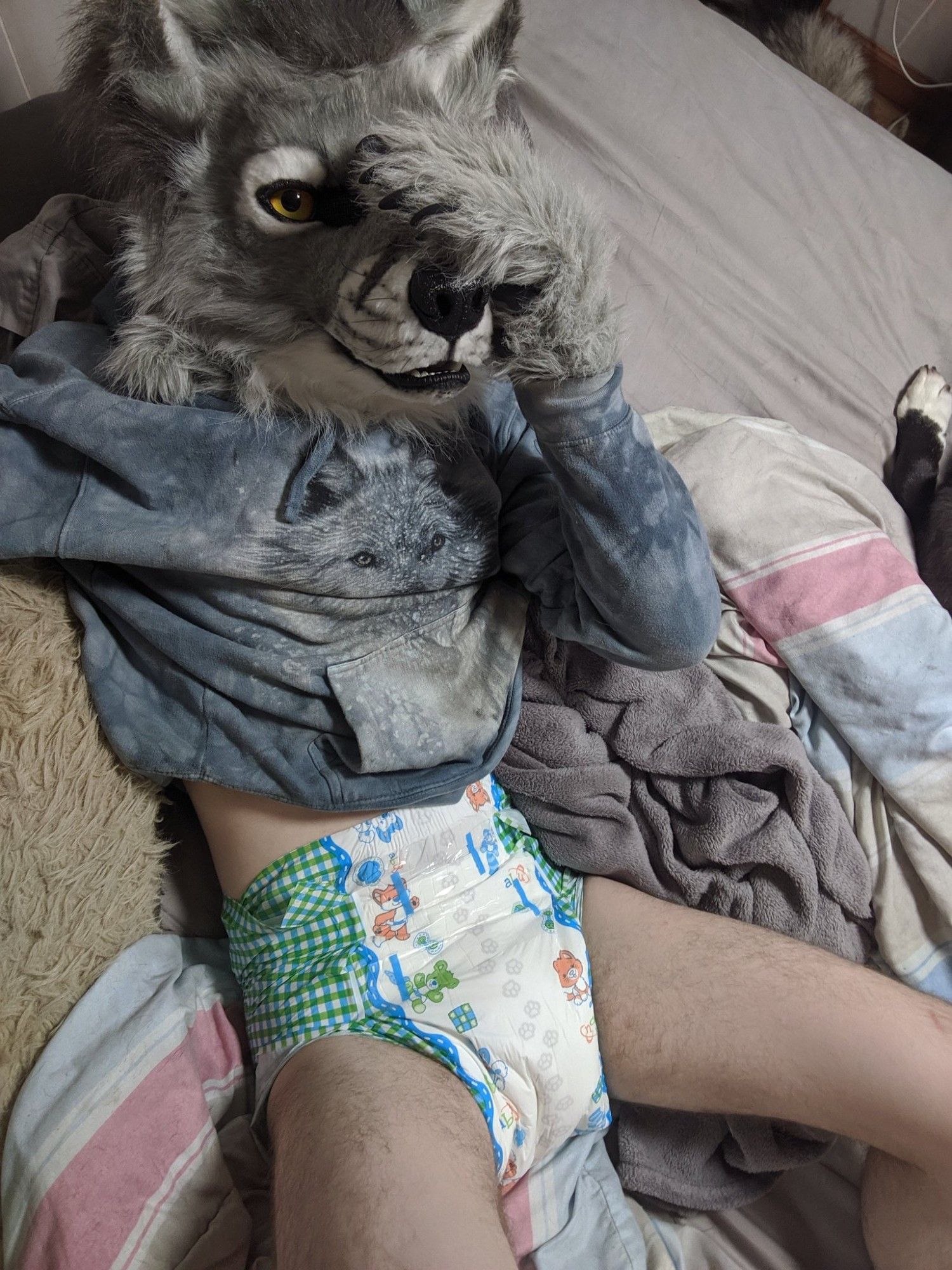 An adult person laying on their back and wearing a hooded sweatshirt, wolf fursuit head, and disposable "littlepawz" diapers.