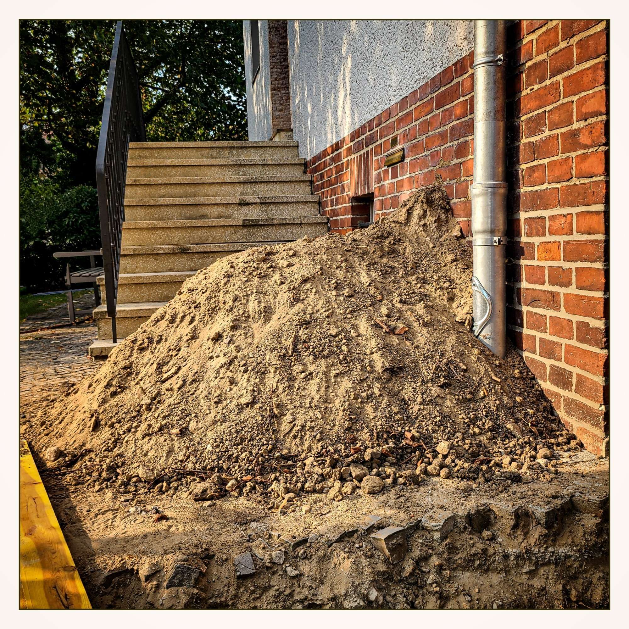 Größer  Kieshaufen, der sich direkt neben einer Treppe und einer Hauswand aus roten Ziegeln befindet. Die Treppe führt zu einem Eingang, und an der Hauswand ist ein silbernes Fallrohr zu sehen. Es scheint, als ob hier Bau- oder Grabungsarbeiten stattfinden.