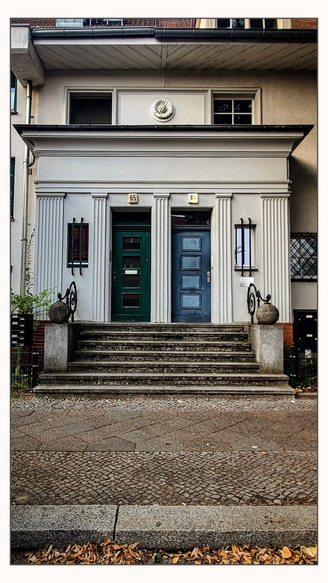 Eingang eines Gebäudes mit zwei nebeneinanderliegenden schlanken Türen, die durch eine breite Treppe erreichbar sind. Eine Tür ist grün, die andere blau. Über den Türen sind die Hausnummern 65 und 64 angebracht. Die Architektur ist klassizistisch geprägt, mit Säulen und einem dekorativen Relief über den Türen.