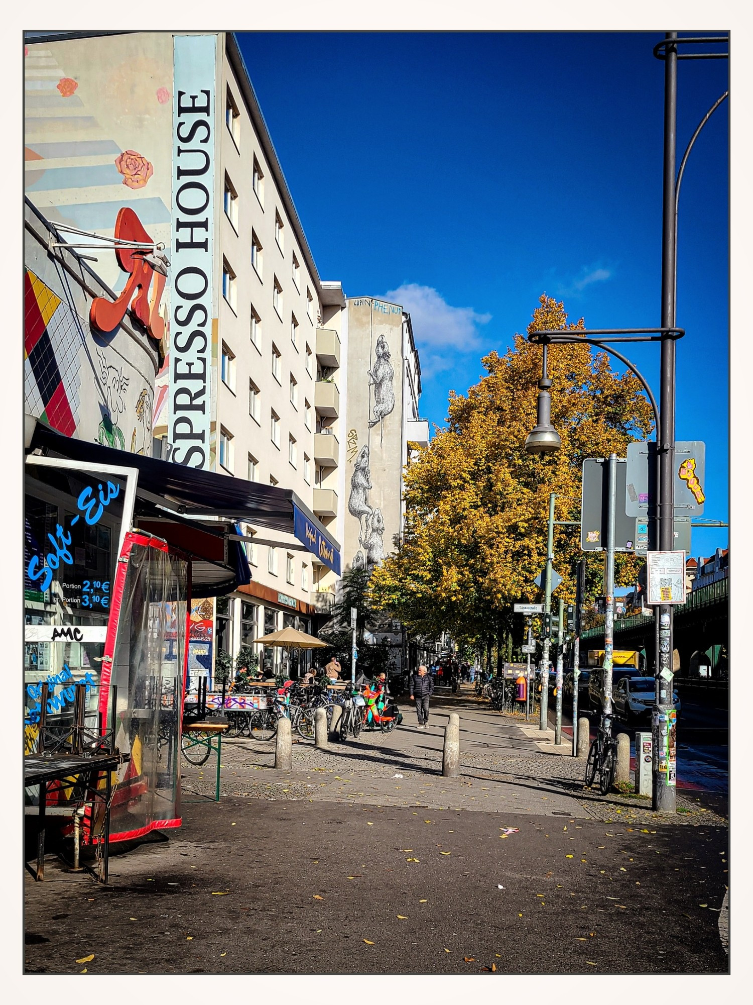 Straßenszene bei sonnigem Wetter und strahlend blauem Himmel. Links ist ein Gebäude mit der Aufschrift „Espresso House“ zu sehen, und es gibt mehrere bunte Graffitis an den Wänden. Im Vordergrund befindet sich ein Imbiss mit umgedrehten Bierbänken davor und einer ausgezogenen Markise. Weiter hinten einige Fahrräder. Rechts säumen Bäume mit herbstlichem Laub die Straße. Menschen in der Ferne spazieren entlang des Gehwegs.