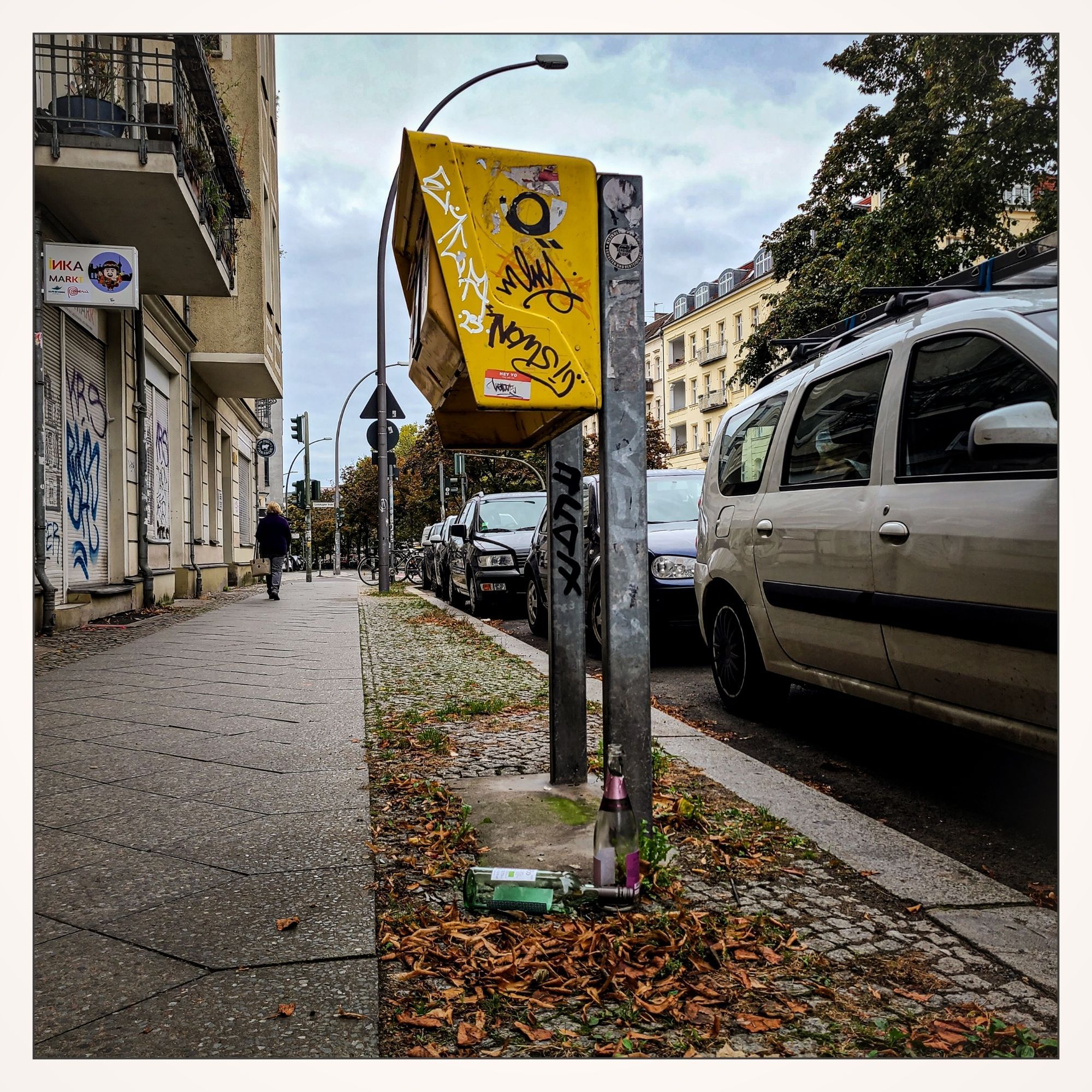 Blick auf zwei Weinflaschen unter einem gelben Postbriekaste. Eine helle Flasche steht, eine grüne liegt daneben.
Rechts parkende Fahrzeuge. Links der Gehweg mit angrenzenden Häusern.