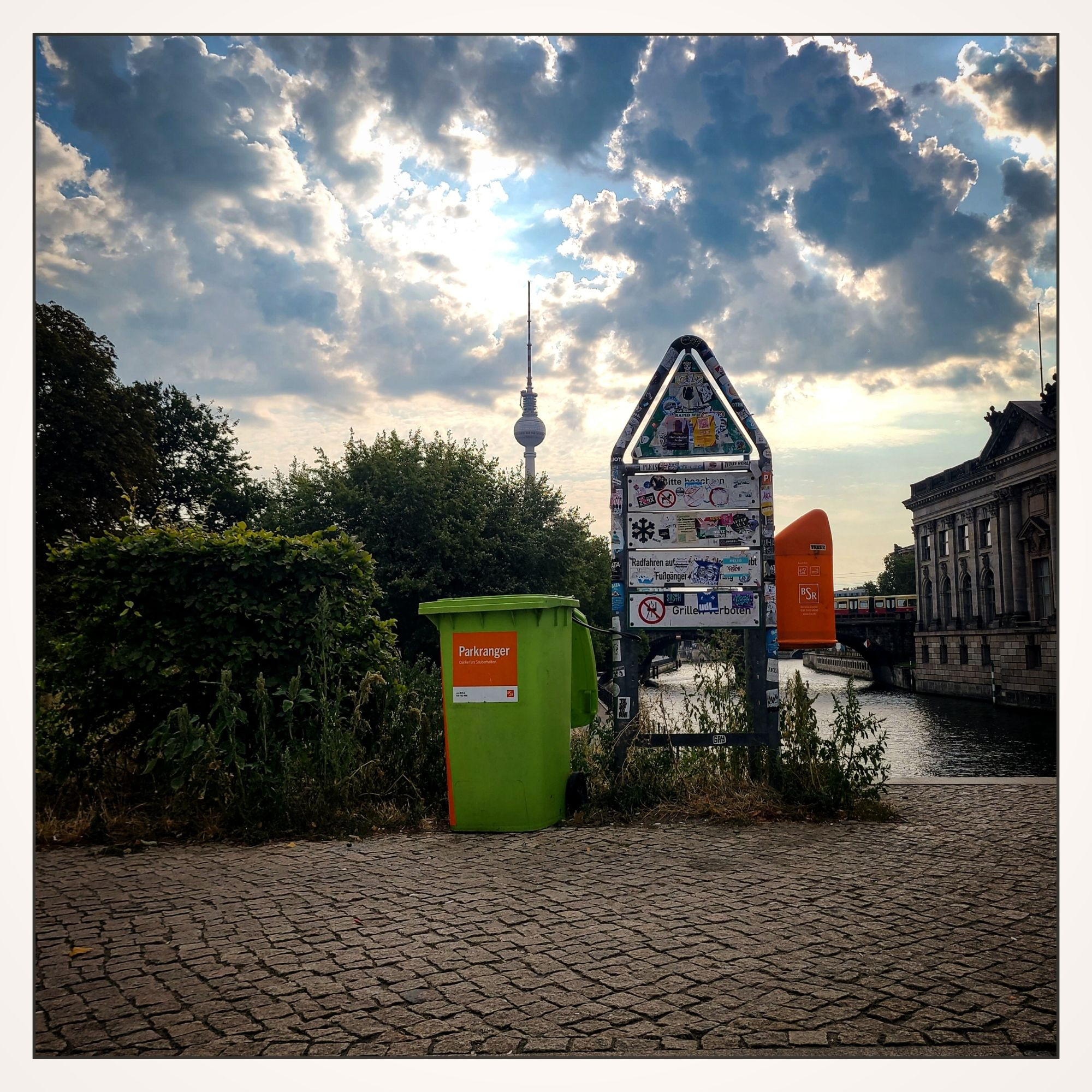 Städtische Szene mit einer mit Aufklebern bedeckten Informationstafel, einer kleinen grünen Mülltonne mit der Aufschrift "Parkranger" und einem orangen Mülleimer. Im Hintergrund ist der Berliner Fernsehturm zu sehen, umgeben von Bäumen und einem Fluss, über den eine Brücke führt. Die Wolken am Himmel sind dicht, aber die Sonne scheint hindurch.