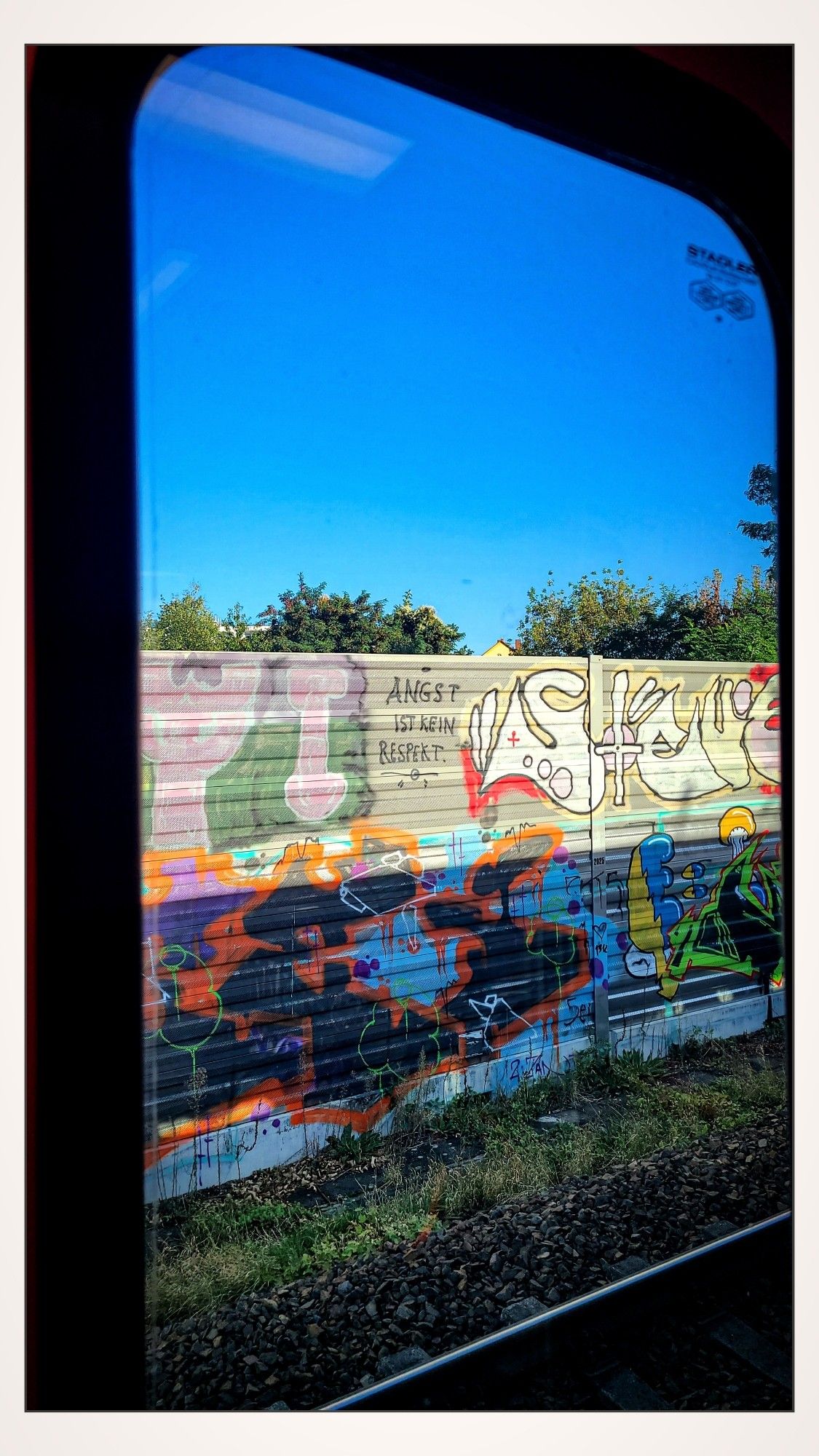 Blick aus einem stehenden Zug auf eine graue Geräuschschutzwand, die voller bunter Graffiti ist. Dazwischen der Spruch "Angst ist kein Respekt". 
In der rechten unteren Ecke sieht man ein Teil des Gleises. Über der Wand kommt strahlend blauer Himmel zum Vorschein.