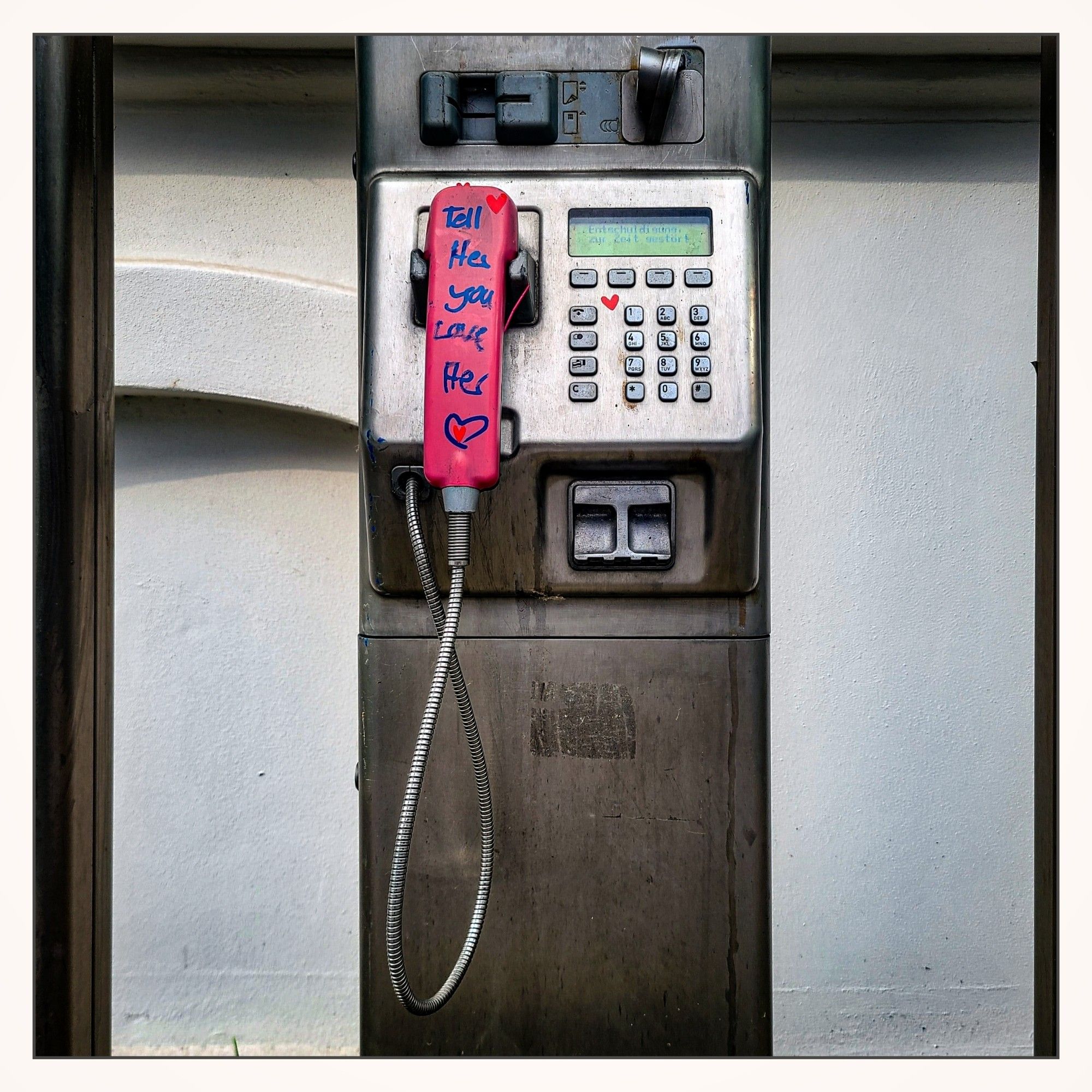 Alte offene Telefonstele der Telekom. Der Apparat hat einen Münzeinwurf, ein Tastenfeld und an der intakten Telefonstrippe aus Metall ist der pinke Hörer eingehängt. Auf diesem steht geschrieben: Tell her you love her <3.