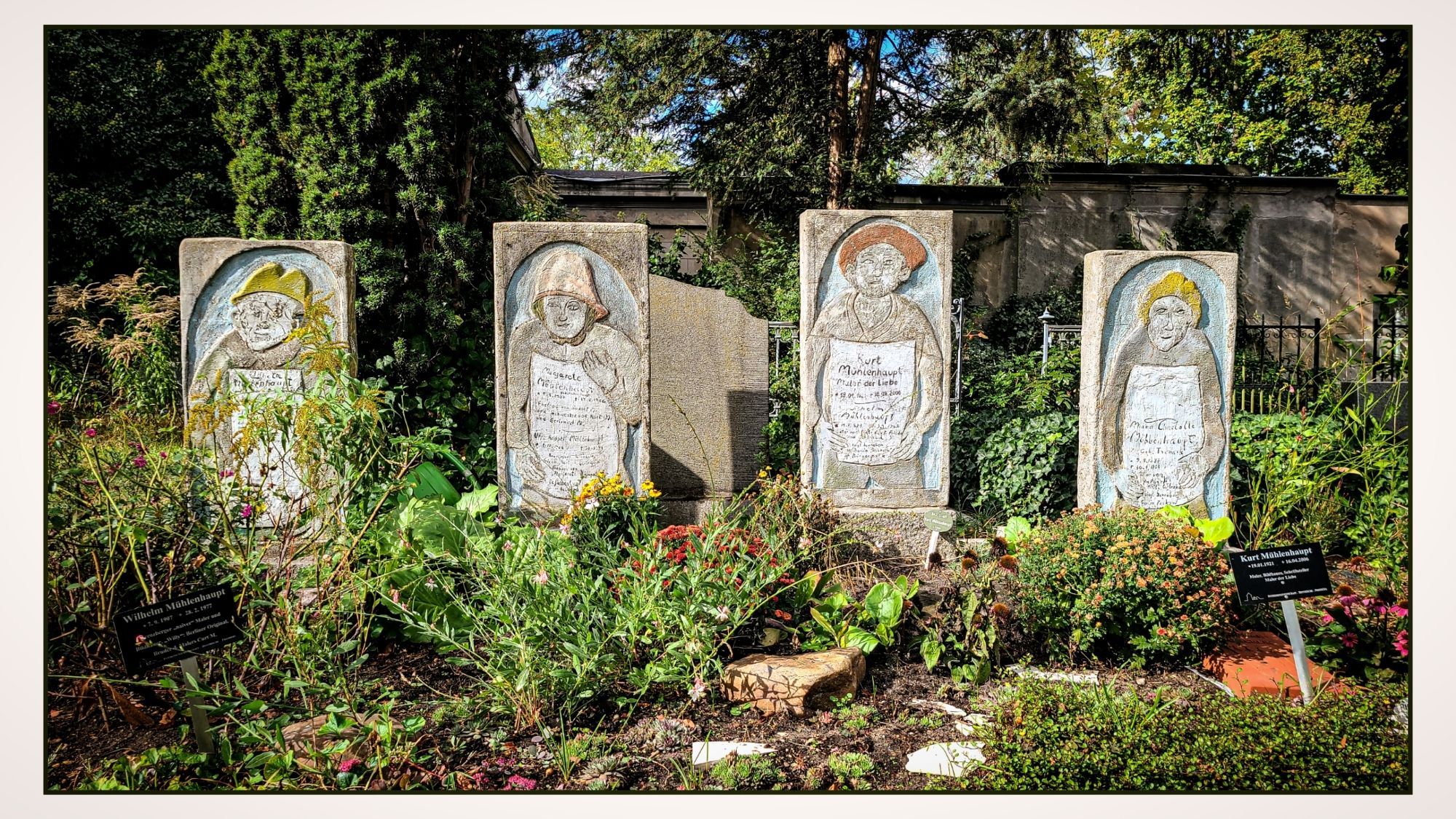 Vier aufrecht stehende Grabsteine nebeneinander auf einem Friedhof. Auf jedem Stein ist eine Figur als Relief abgebildet, sowie eine Inschrift darunter. Die Gruppe gehört quasi als Familiengrab zusammen. 
Der Bereich ist von üppiger Vegetation und Blumen umgeben. Auf zwei kleinen Tafeln im Vordergrund sind die Namen "Wilhelm Mühlenhaupt" und "Kurt Mühlenhaupt" zu lesen, die offenbar mit den dargestellten Personen in Verbindung stehen.