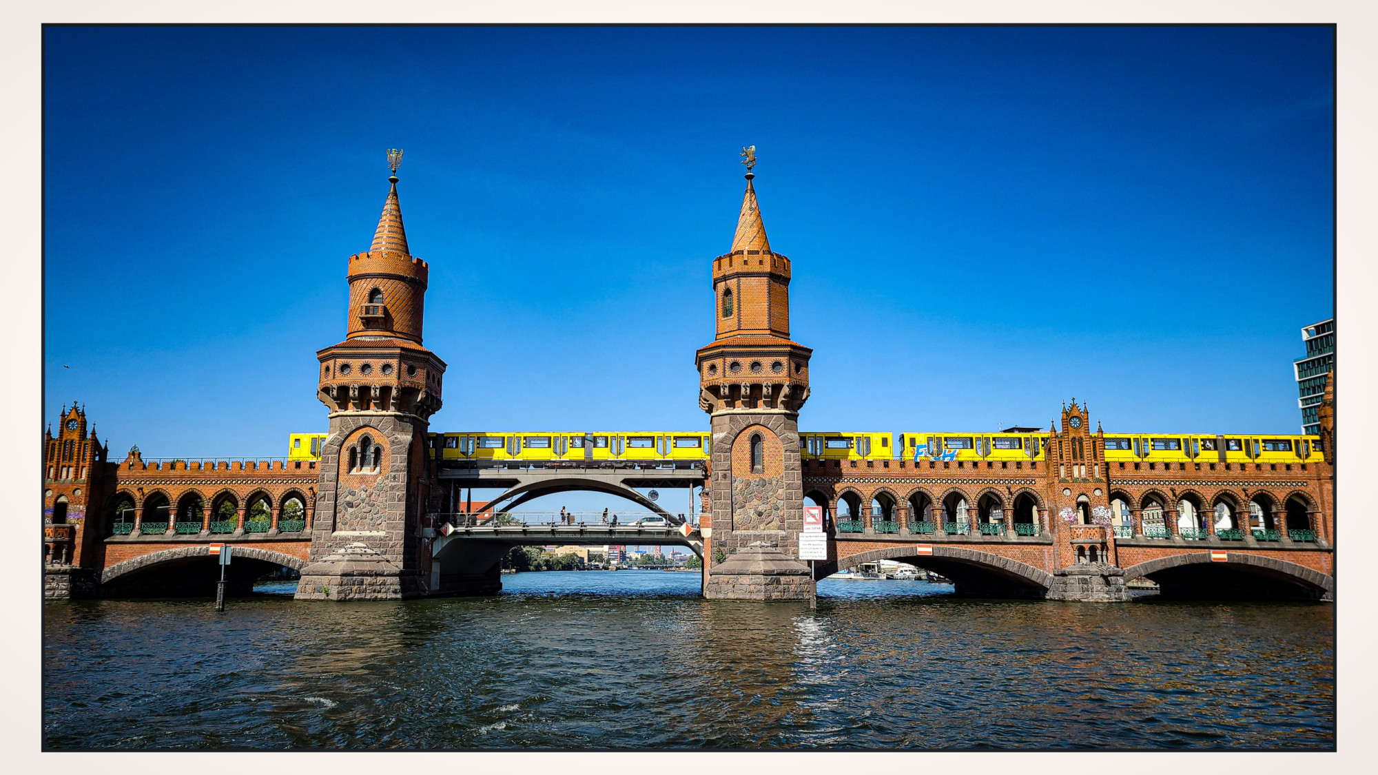 Eine über dem Wasser verlaufende Brücke aus Backstein mit zwei hübschen verzierten Spitztürmen. Eine gelbe U-Bahn fährt dort gerade entlang. Strahlend blauer Himmel.