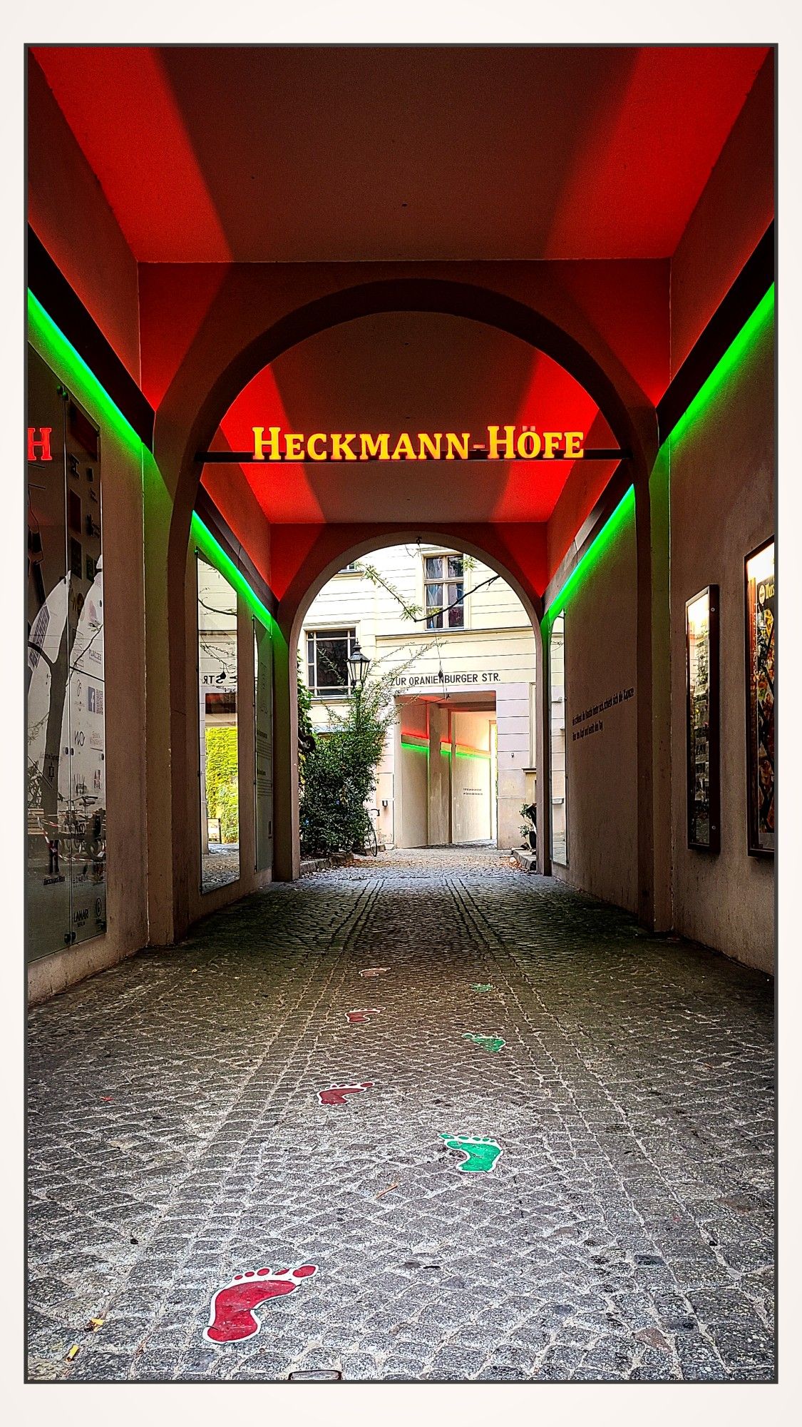 Hausdurchgang mit einem beleuchteten Schild, auf dem „Heckmann-Höfe“ steht. Der Durchgang ist von roten und grünen Neonlichtern umrahmt, und auf dem gepflasterten Boden sind farbige Fußabdrücke (rot und grün) zu sehen, die den Weg weisen. Am Ende des Durchgangs ist ein weiterer Torbogen, der zur Oranienburger Straße führt.

