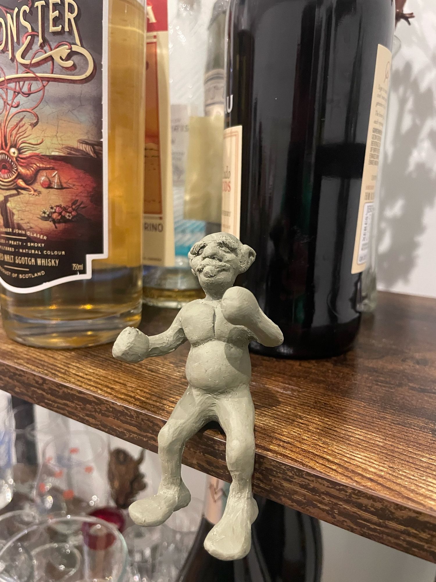 A clay sculpture of a small dwarf or gnome posing among some licquor bottles