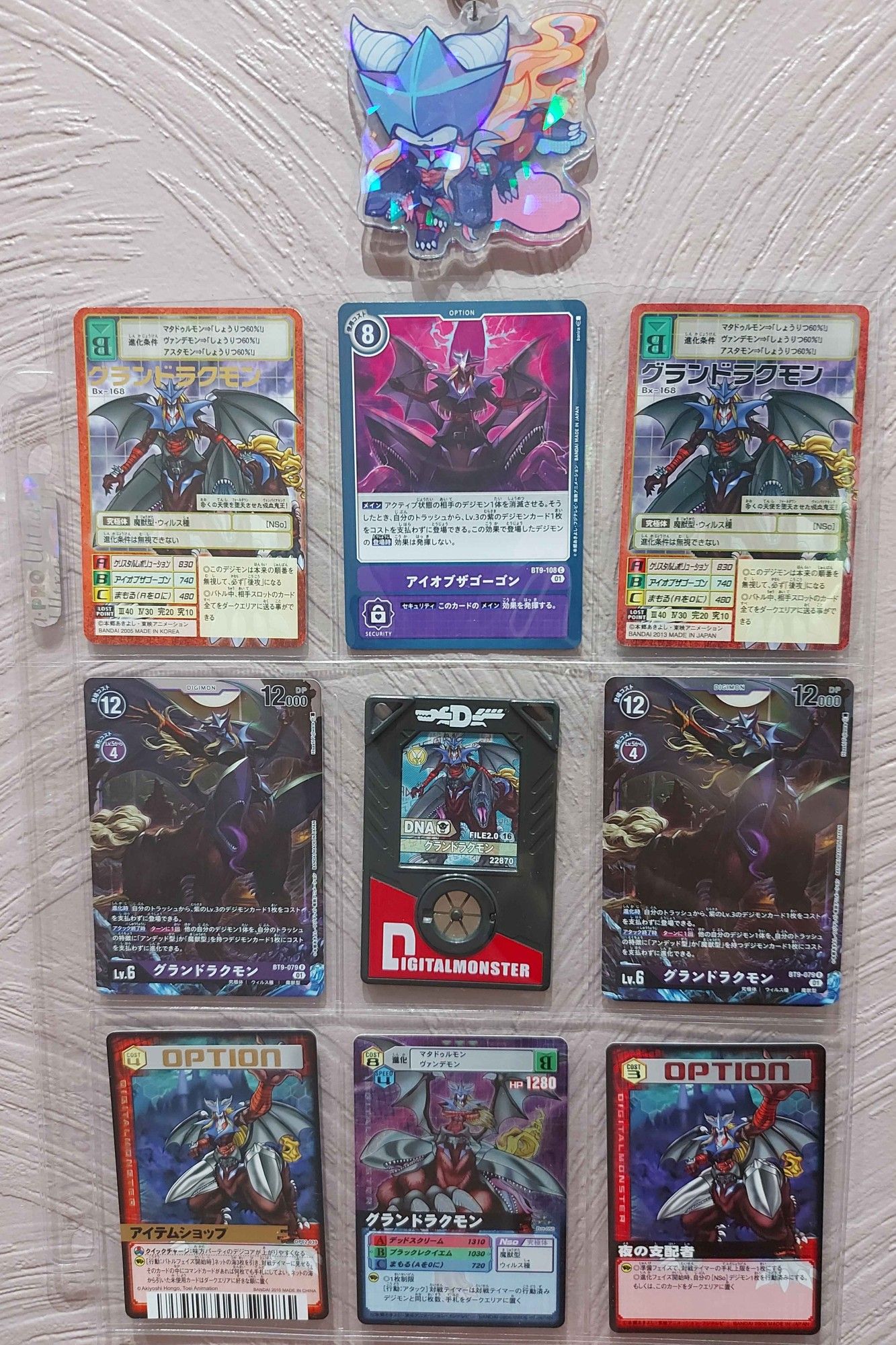 A photo of 9 digimon (GranDracmon) cards in a page protector. Above them is a holographic shiny acrylic keychain of GranDracmon in a chibi art style.