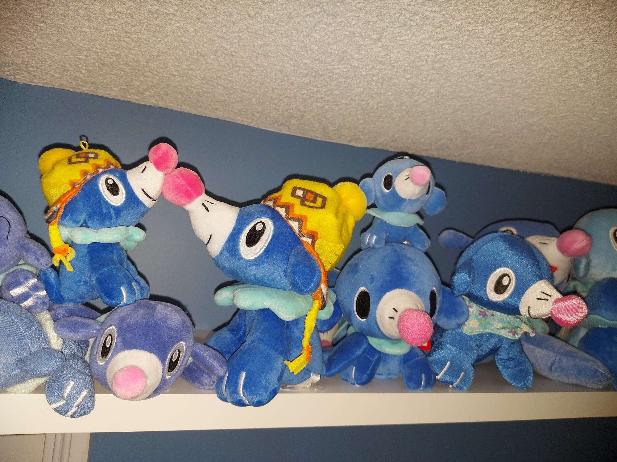 A white shelf over a closet door on a blue painted wall. The shelf is full of pokemon (Popplio) plushies. This photo shows a close up of a few of them, including 2 wearing touques bumping noses.