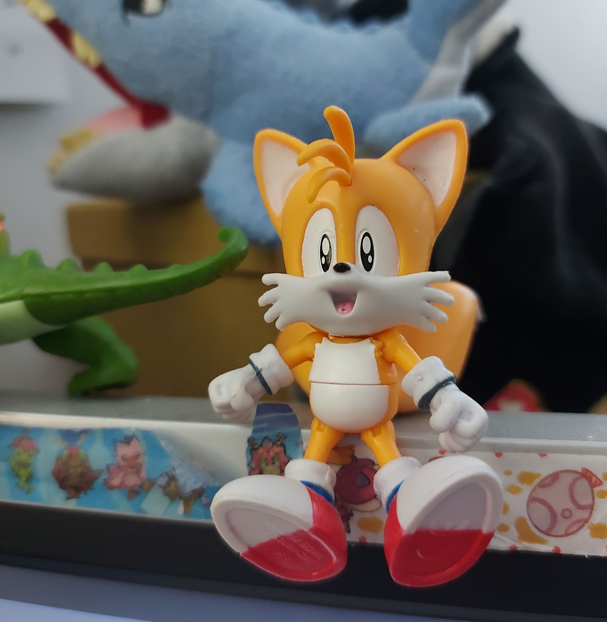 A Tails (from Sonic the Hedgehog) figure sitting on top of a monitor. Other toys and plushies are visible in the background and surroundings.