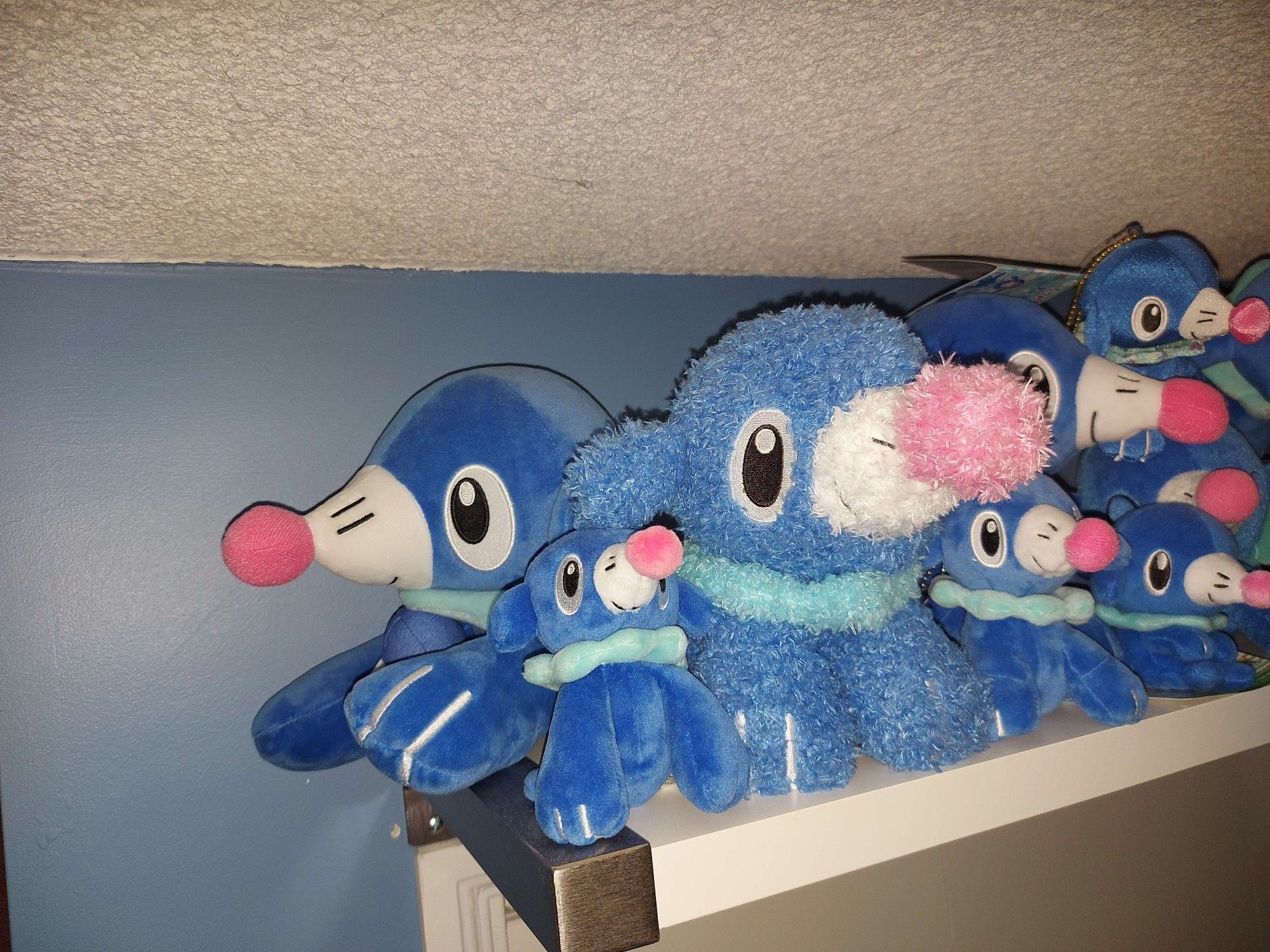 A white shelf over a closet door on a blue painted wall. The shelf is full of pokemon (Popplio) plushies. This photo is a close up of one end of the shelf.