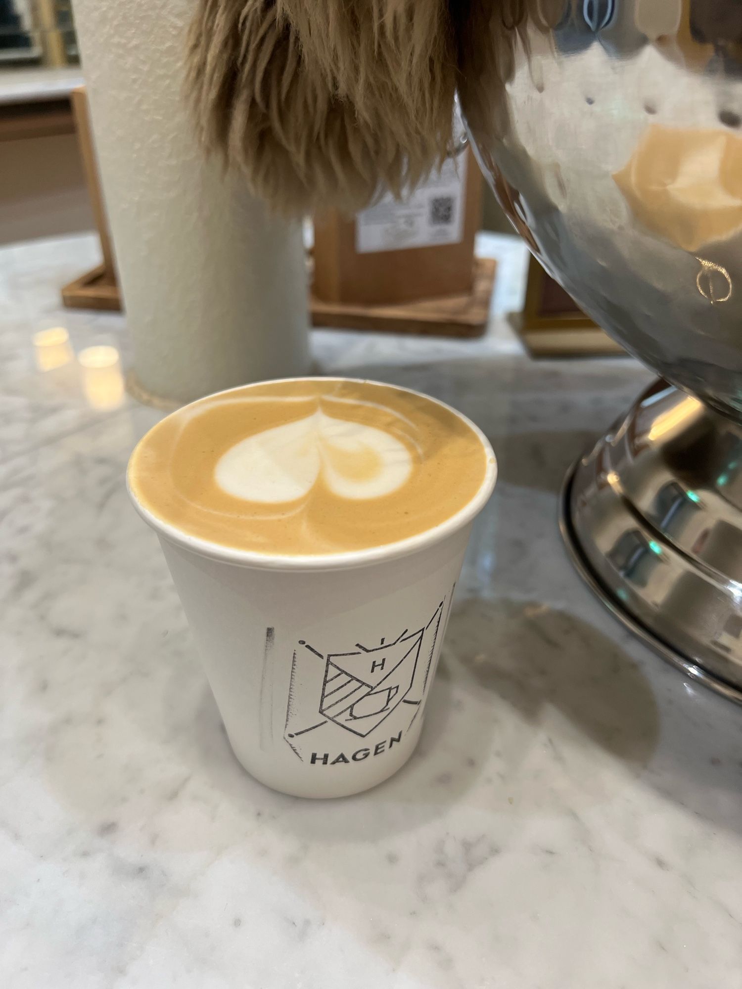 A photo of an oat latte in a white takeaway cup. It has a Hagen branding with a shield logo. There is heart shaped latte art. It sits on a white marble surface. There is a big shiny bowl to the right, reflecting the latte.