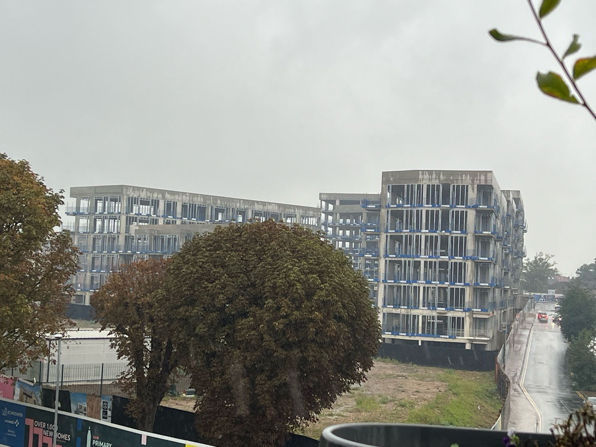 A photo of wet concrete skeletons of buildings. An abandoned construction site. There are trees too, and a road.