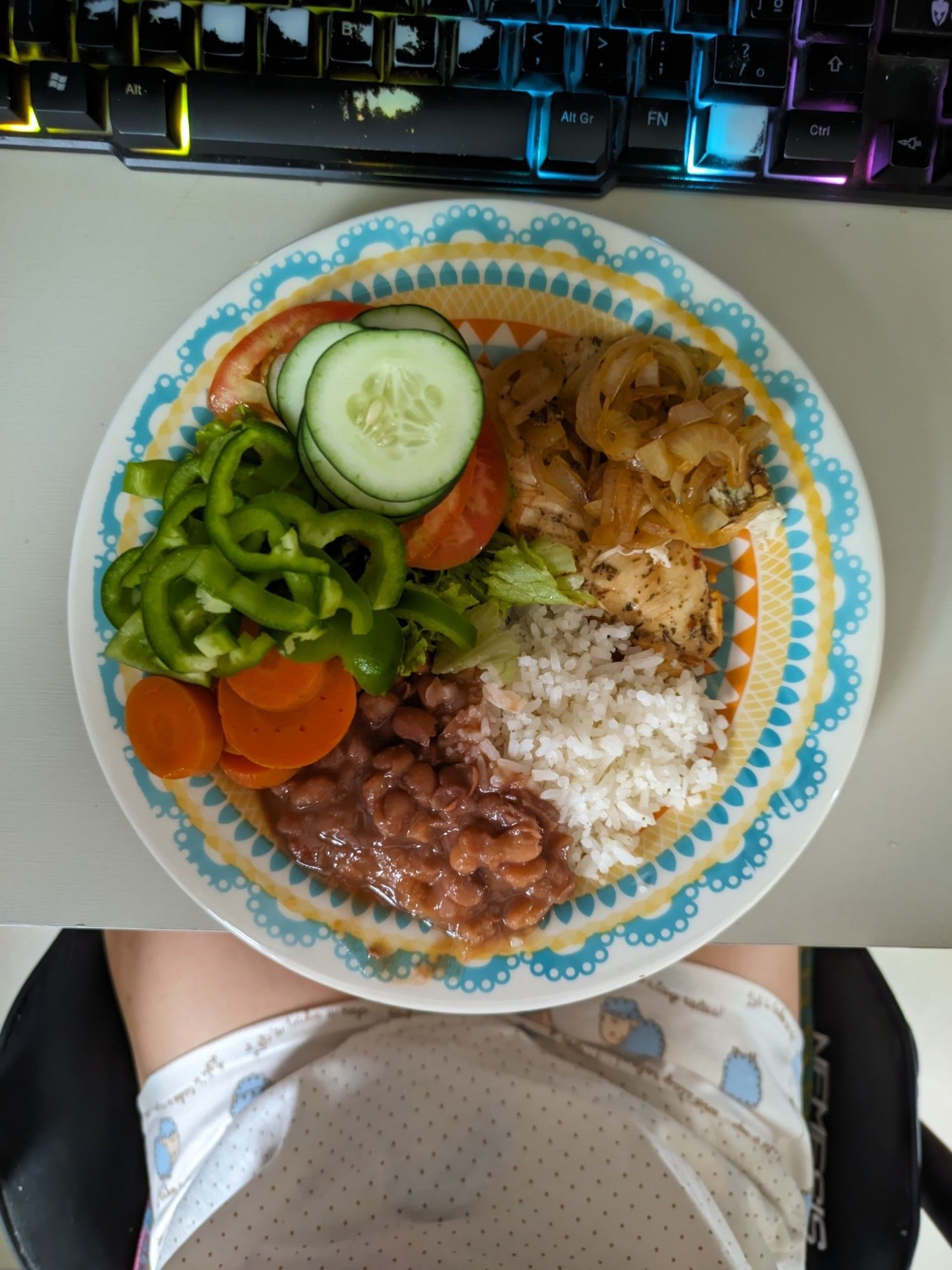 Foto de prato de almoço. A metade esquerda do prato é salda, com alface, rodelas de tomate, rodelas de pepino, pimentão verde picado e rodelas de cenoura cozida. O canto superior direito tem frango acebolado, e o restante é arroz branco com feijão carioca por cima/ao lado.