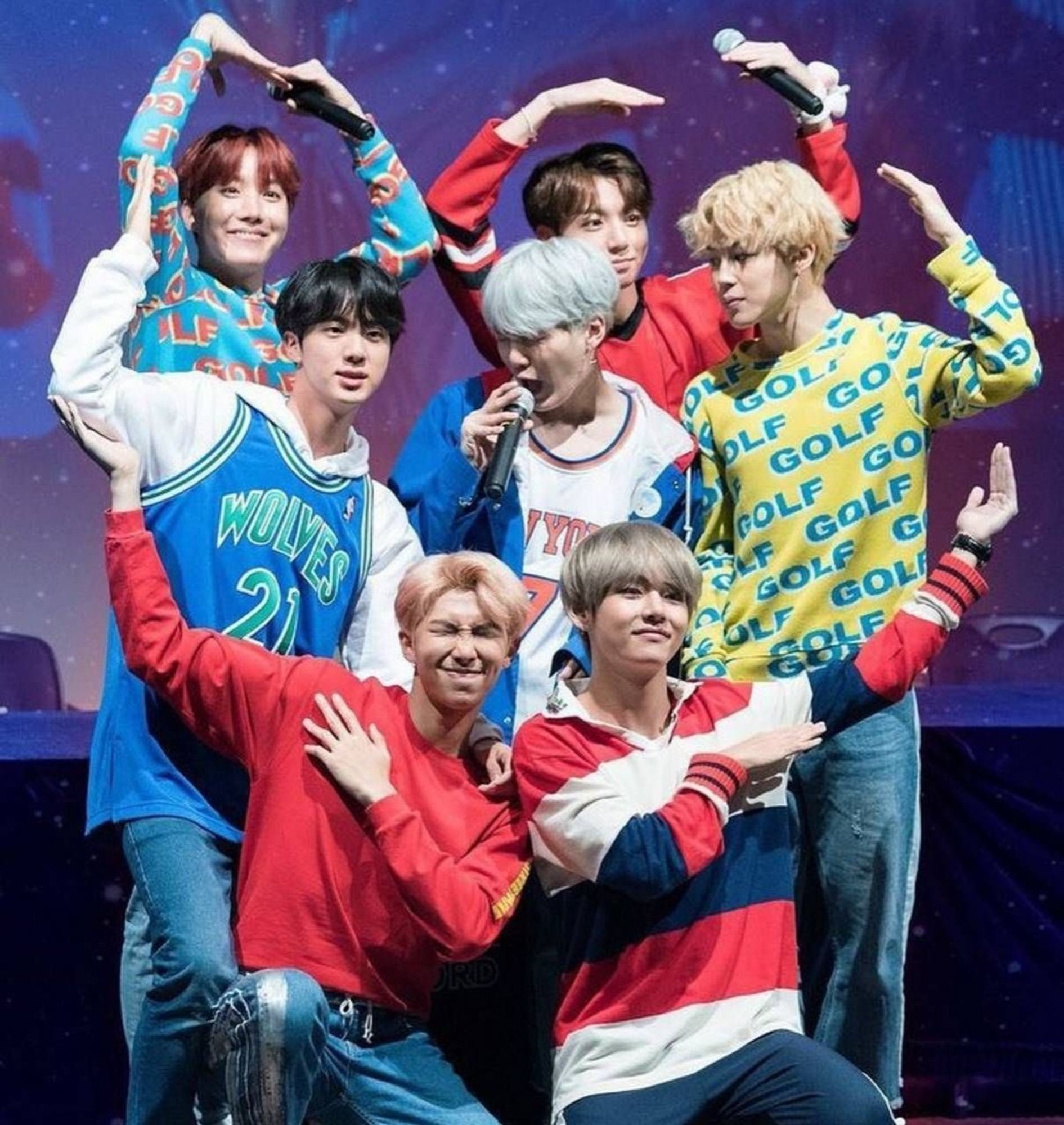 All seven members of BTS on stage, forming a heart with their arms. Dressed in colourful clothes, mainly red and blue.