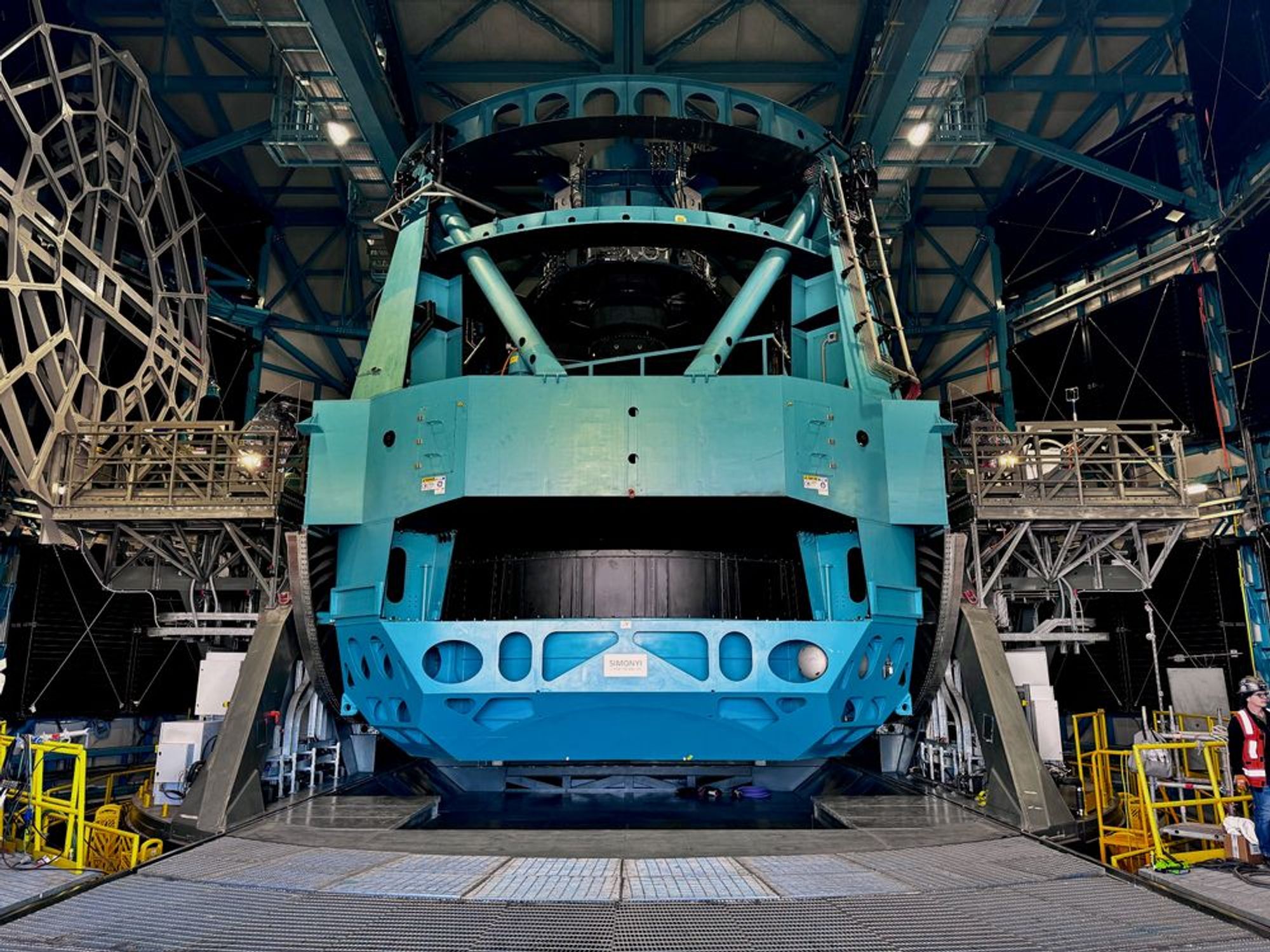 Rubin's telescope structure, pointing upward. The telescope is made of teal steel support beams and is shaped like a vase, with a flat top and rounded flat bottom.