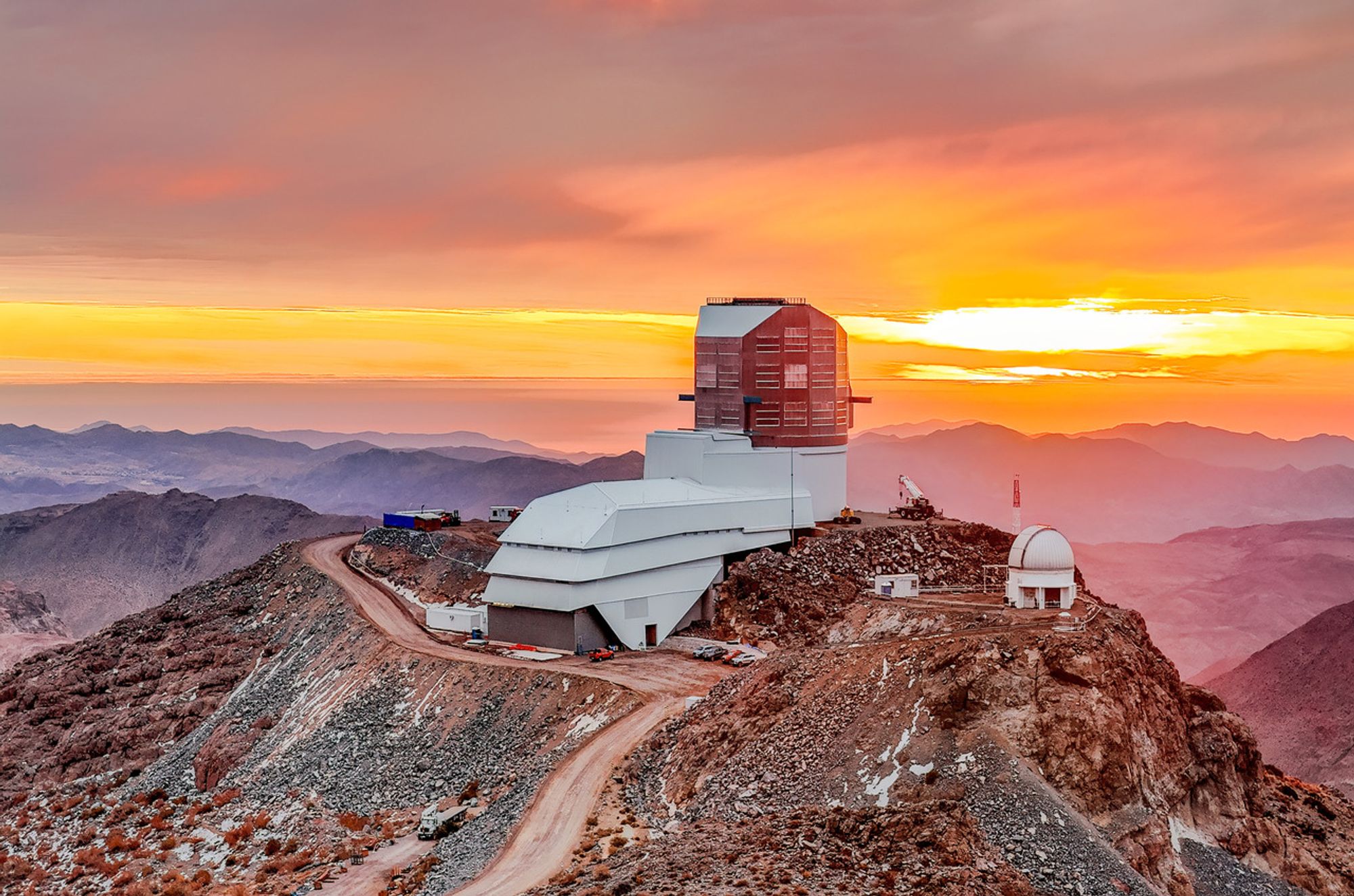 Drone view of Rubin Observatory on top of its summit site on Cerro Pachón against a sunset scene. The observatory building is an angular silver dome on top of a long building extending to the left. The observatory sits against a yellow and orange sky and gray clouds, with rolling mountain ridges in varying shades of purple and pink fading into the background.