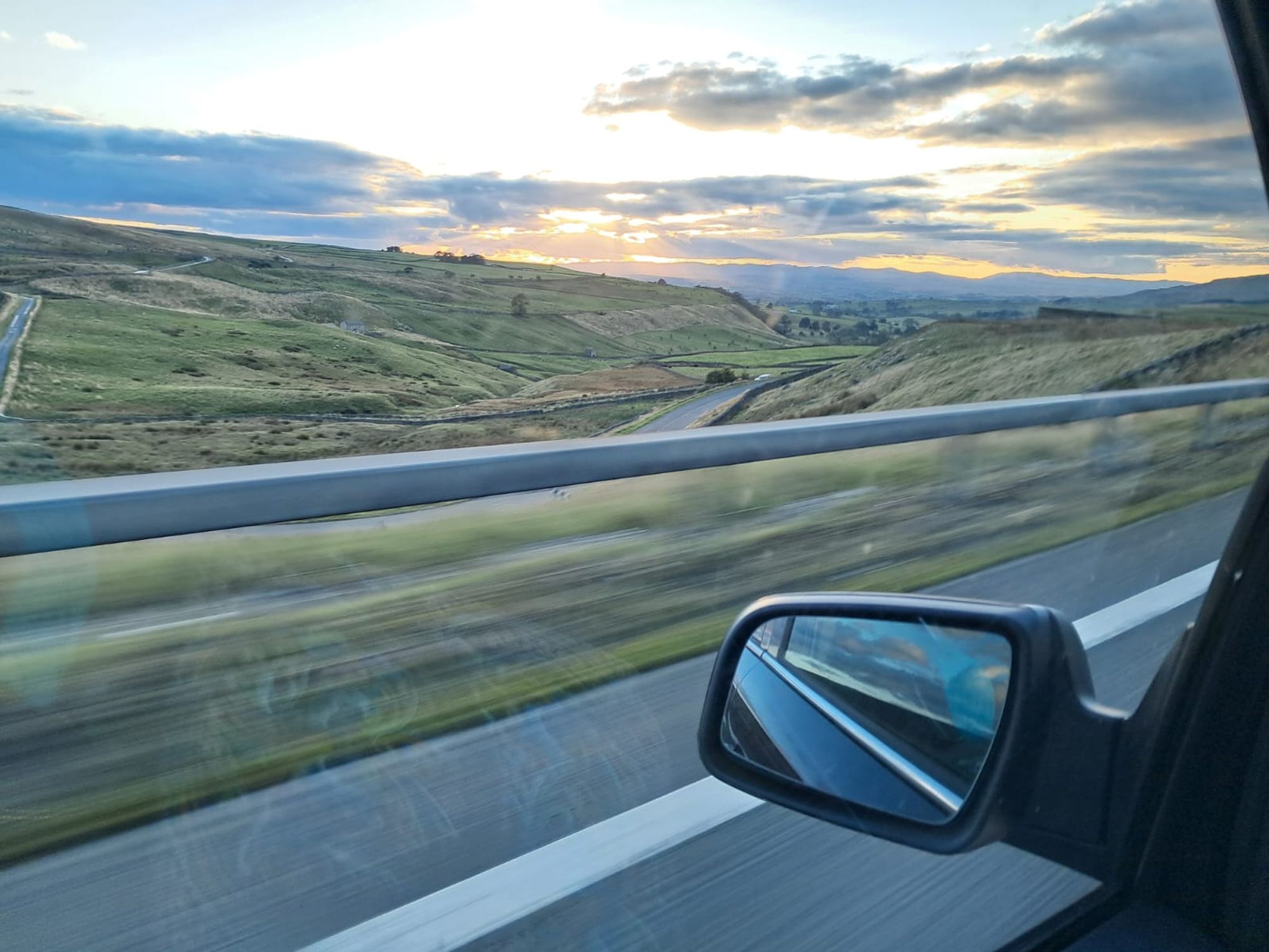 View from the passenger seat of a car. There are hills, country roads and a sunset.