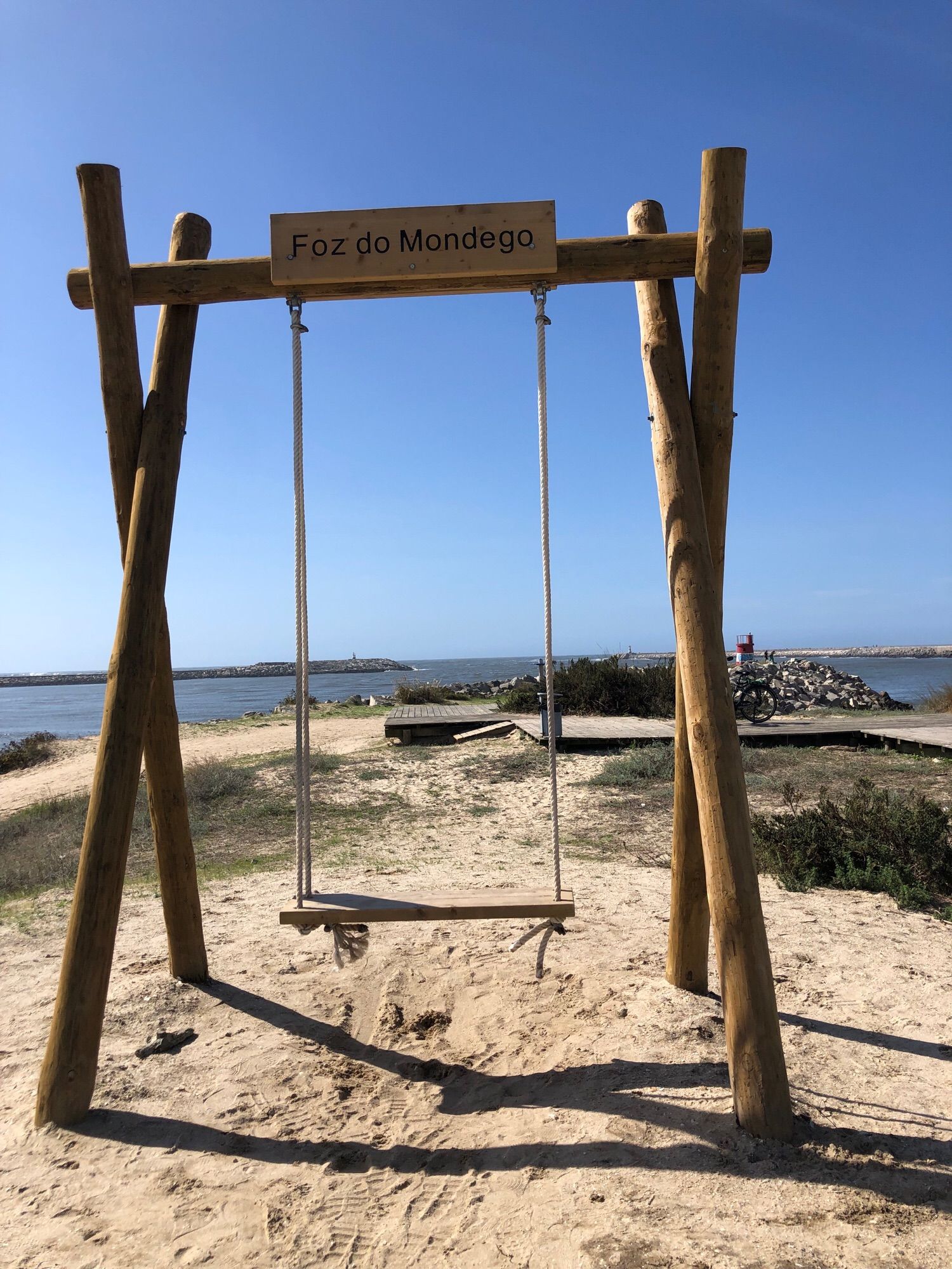 breite, Schaukel, gebaut mit Holzpfählen, Holzbrett zum Sitzen, Holzbrett oben mit der Inschrift „Cabo Mondego. Die Schaukel steht auf einer Sanddüne, im Hintergrund die Fahrrinne für die Schiffe mit Positionsleuchtturm, das offene Meer und darüber blauer Himmel.