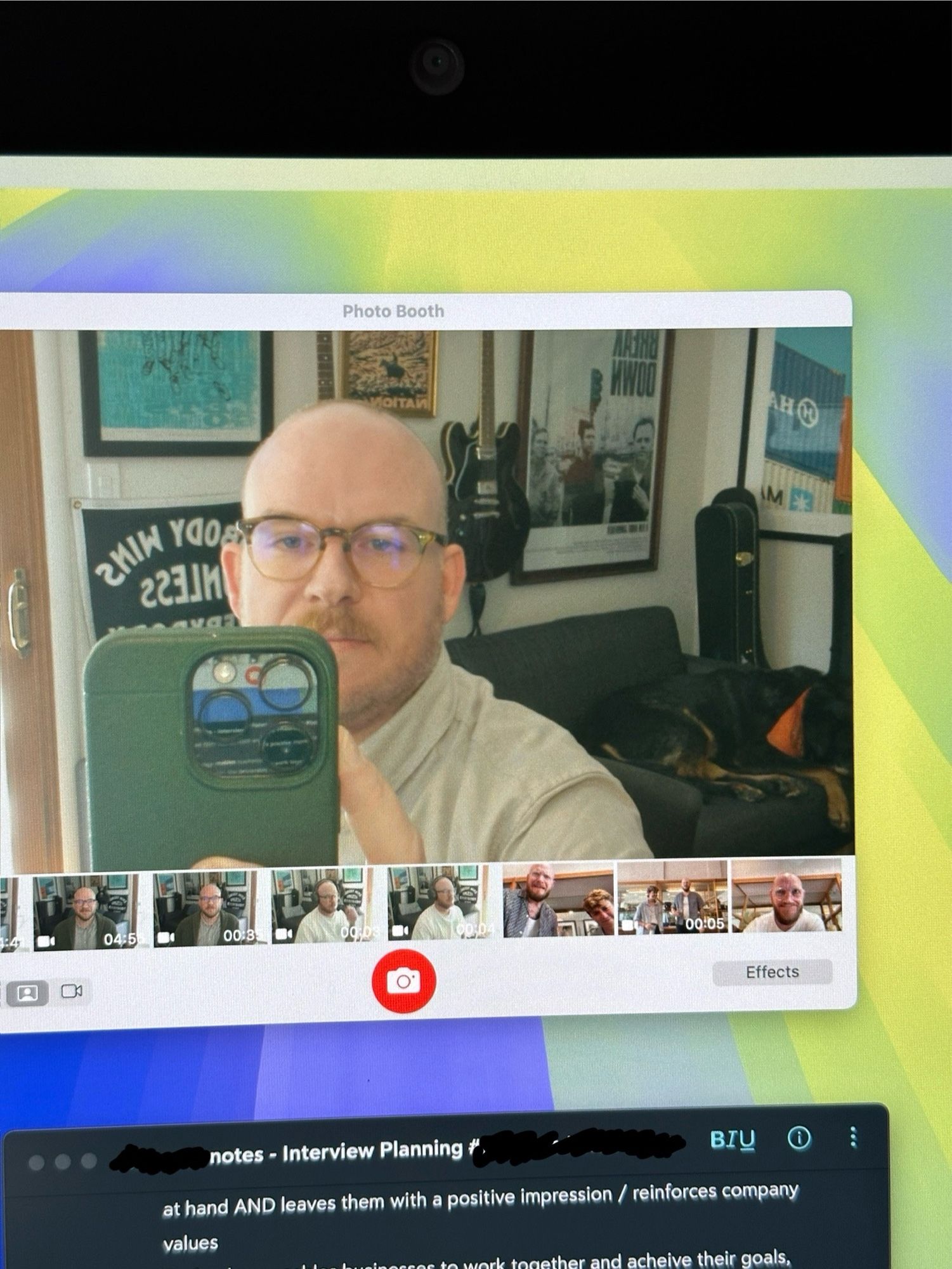 Bald man with glasses takes photo of himself in a camera before an interview. Dog in the background