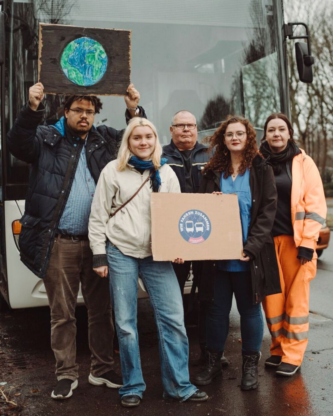 5 Menschen stehen vor einem Bus.  Zu sehen sind: 1 Person of Color mit einem Schild mit einer Erde darauf. Eine Studentin vorne mit einem Schild, wo das Wir Fahren Zusammen Logo drauf ist, daneben ein Busfahrer von der BVG, eine Krankenpflegerin und eine weiblich gelesene Person von der Berliner Stadtreinigung.