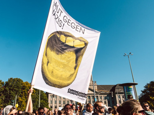 Im Fokus: Eine Fahne auf der Unteilbar Demonstration 2018
Titel laut gegen Nazis: Symbol Mund gelbd eingefärbt, der schreit