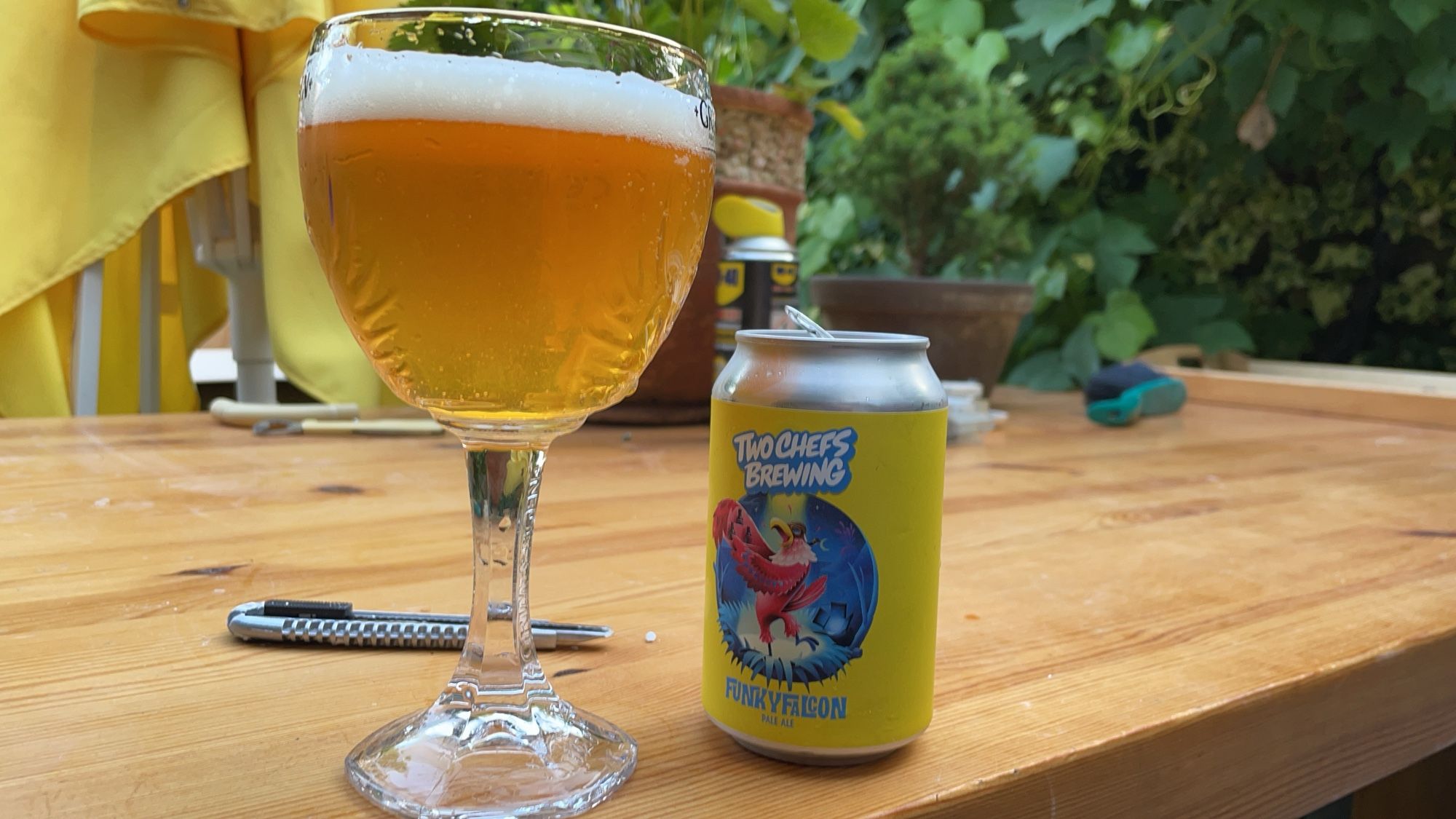 Funky falcon blikje bier met vol glas er naast op een zomerse terrastafel. Toevallig ligt er ook een stanleymesje en een handvegertje bij, details..