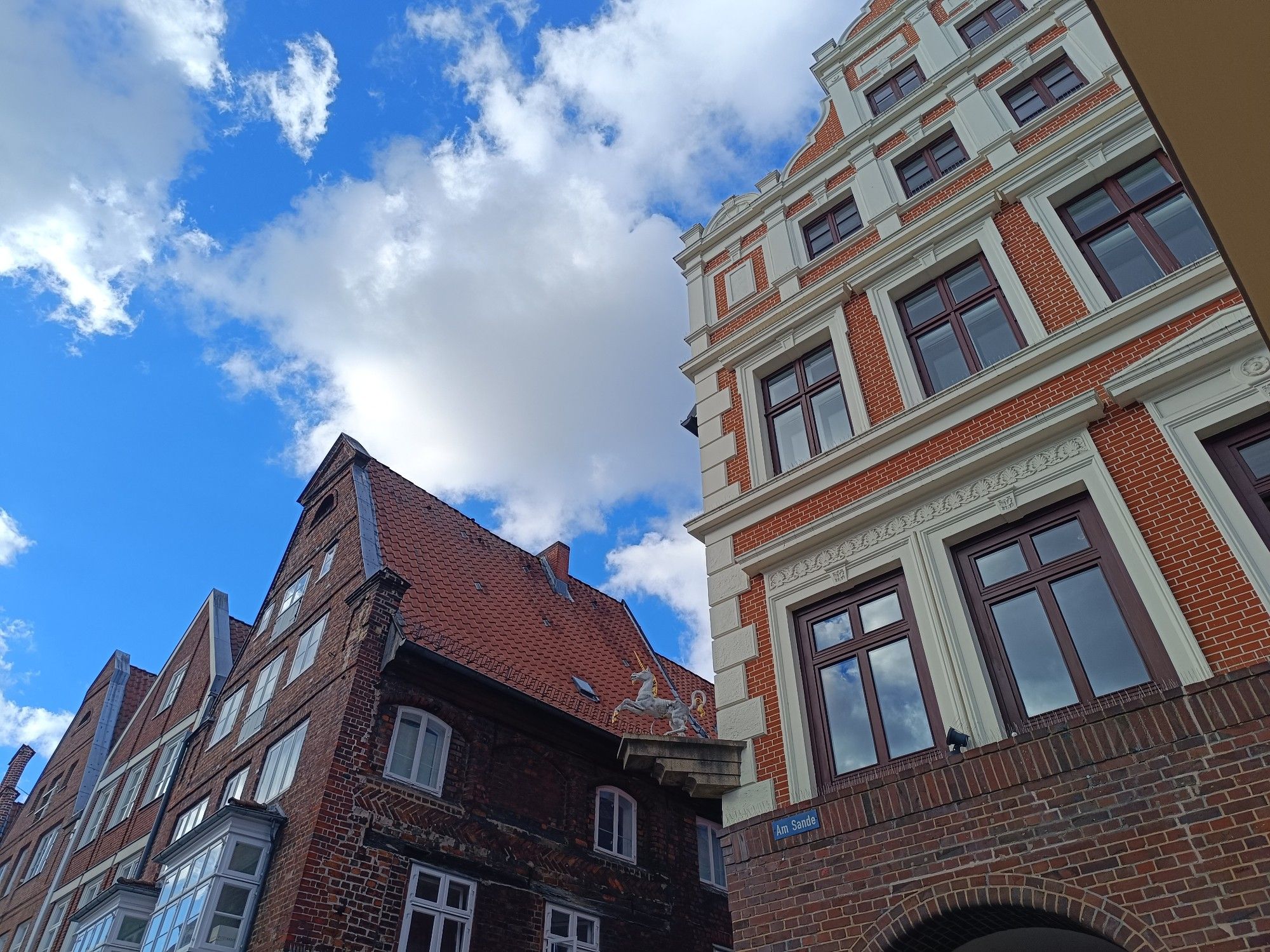 Blick auf zwei Häuser von unten, dazwischen und darüber blauer Himmel, weiße Wolken. Ein Einhorn und Schild "Am Sande".