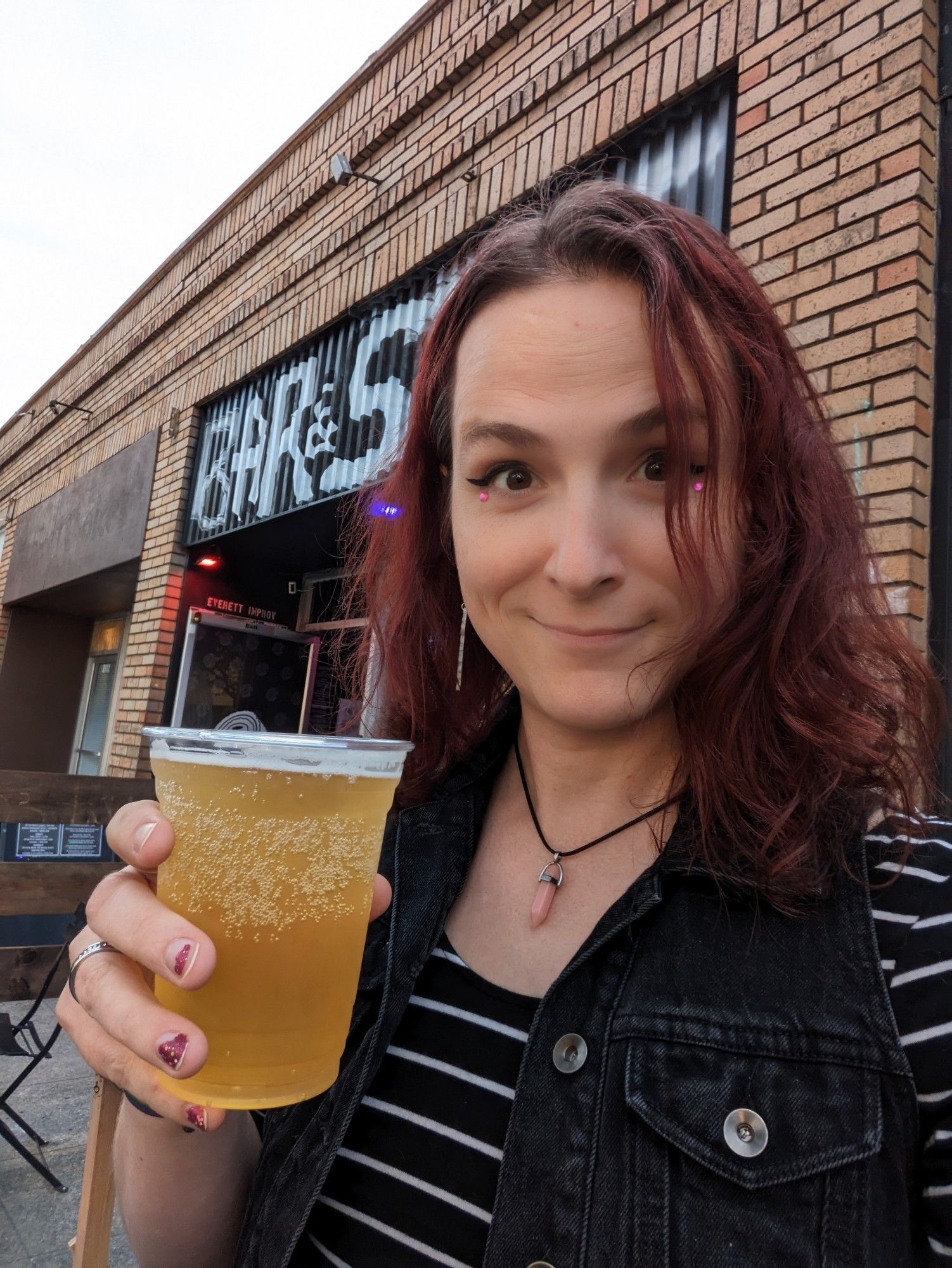 Jacey is wearing a white and black striped shirt and a black vest with cute slightly sparkly makeup, holding a plastic cup of cider up and smiling while sitting outside of a brick building