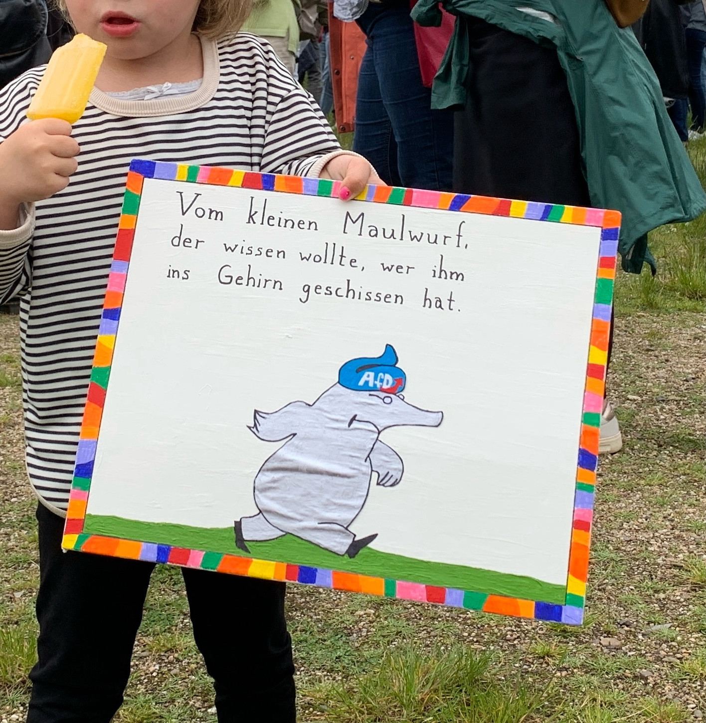 Kind hält ein Schild im Stil des Kinderbuchs „Vom kleinen Maulwurf, der wissen wollte, wer ihm auf den Kopf gemacht hat“ mit dem geänderten Text: „Vom kleinen Maulwurf, der wissen wollte, wer ihm ins Gehirn geschissen hat“. Darunter der Maulwurf mit einem blauen AfD-Kackhäufchen auf dem Kopf.