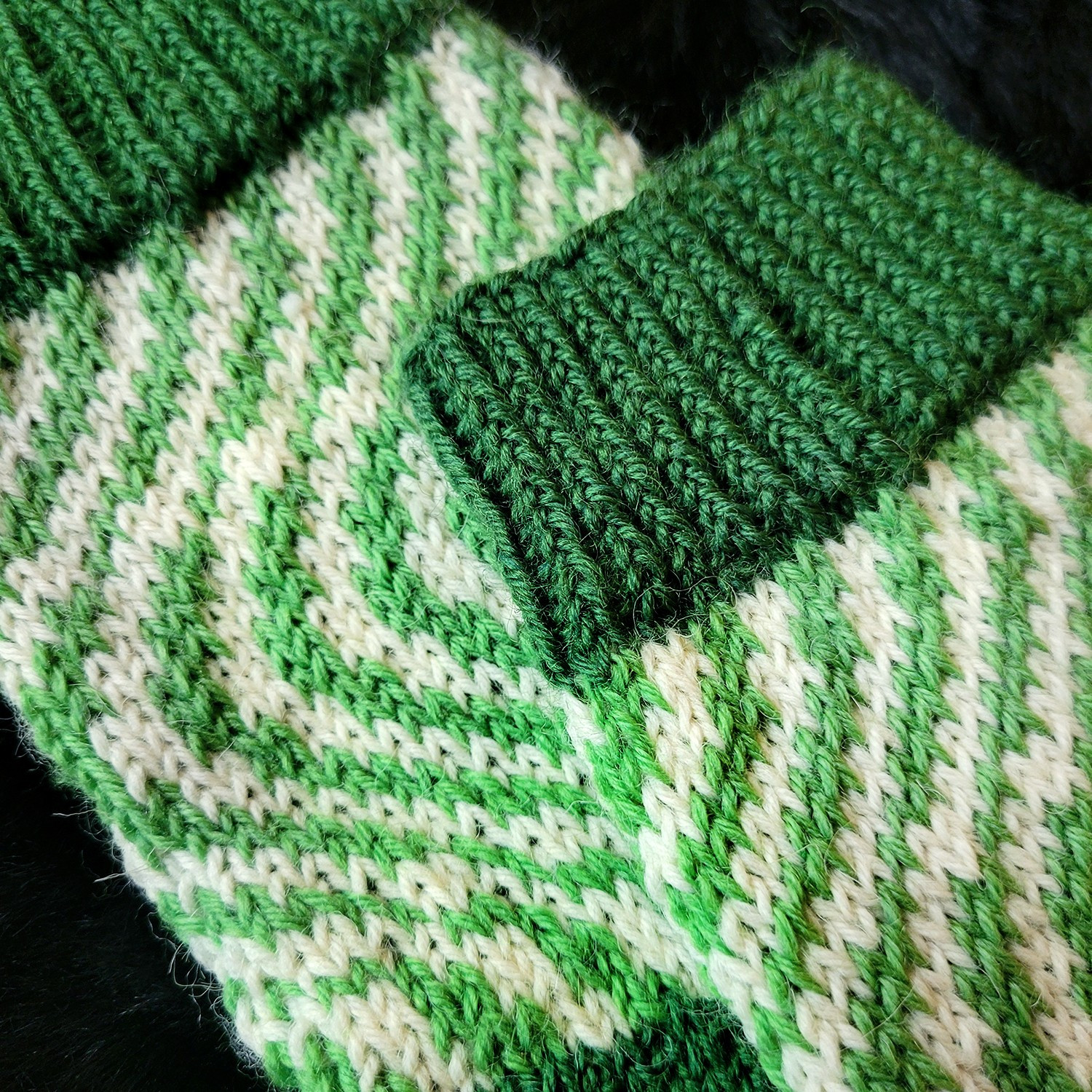 A detail of knitted socks on a black background. The cuff is made in a darker green, while the rest of the socks are draped in lighter green and white abstract colorwork.