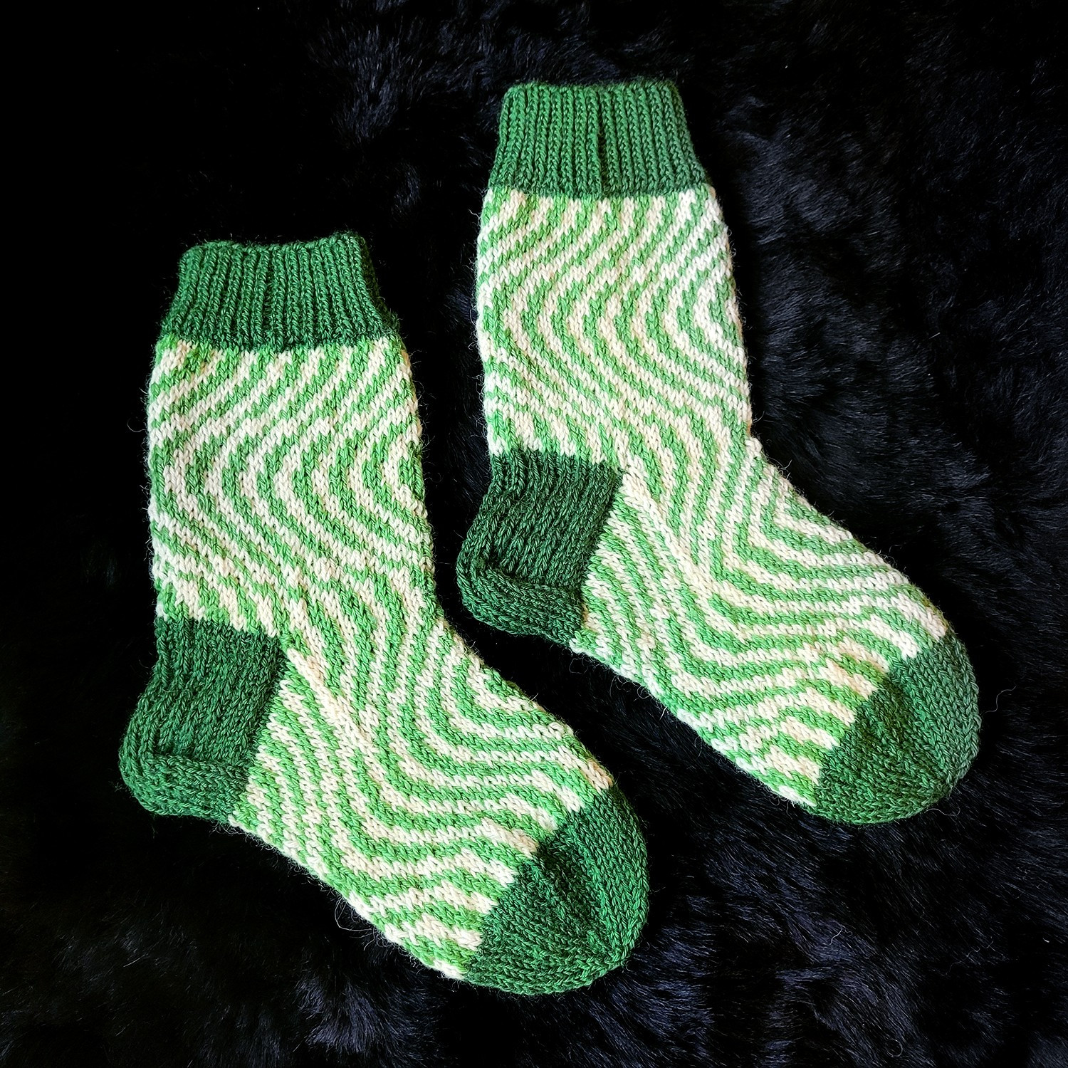 A pair of knitted socks on a black fur background. The heel, toes and cuff are made in a darker green, while the rest of the socks are draped in lighter green and white abstract colorwork.