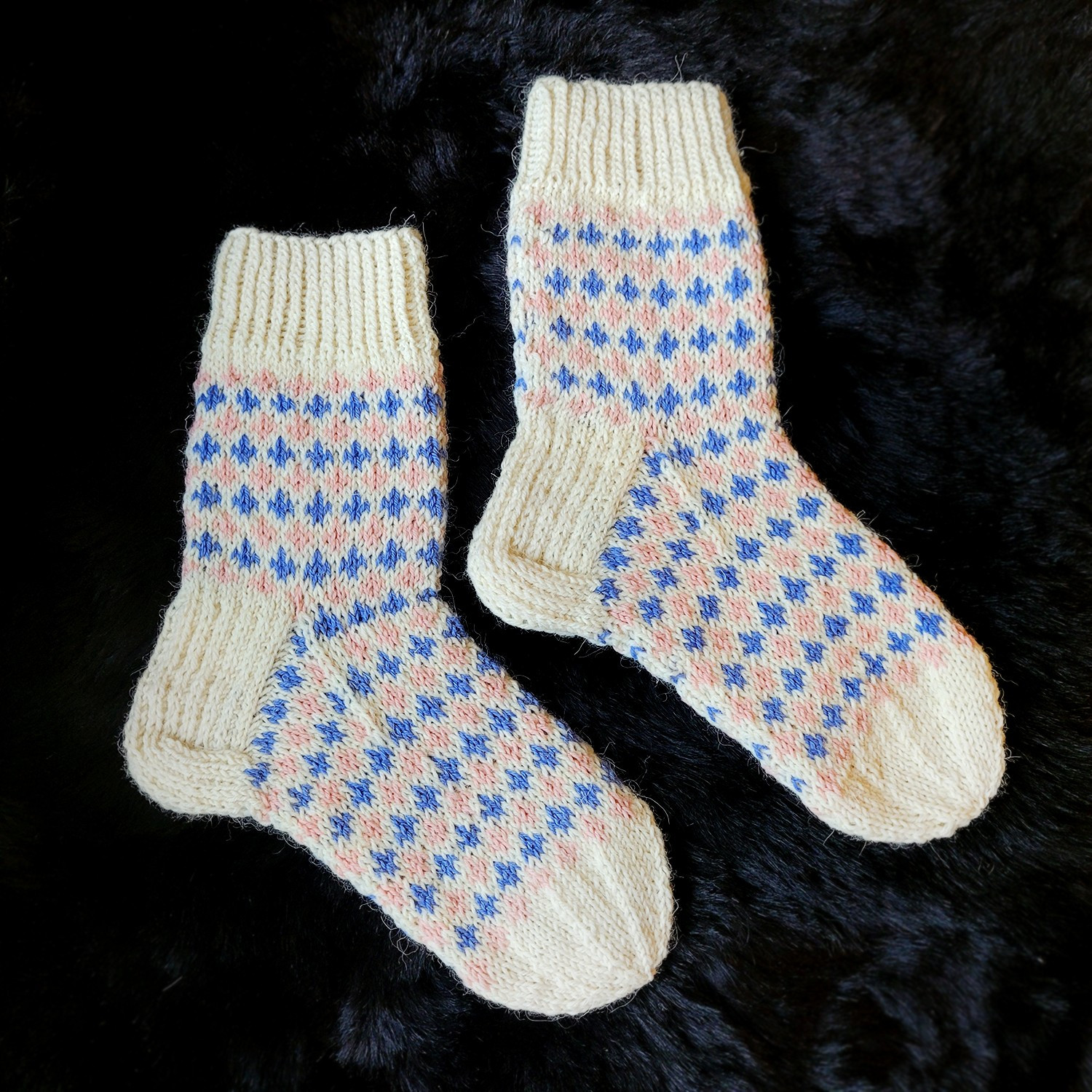 A pair of knitted socks on a black fur background. A pattern of pink and blue fleur de lis extends through the sock, with the exception of the cuff, heel and toe being white.