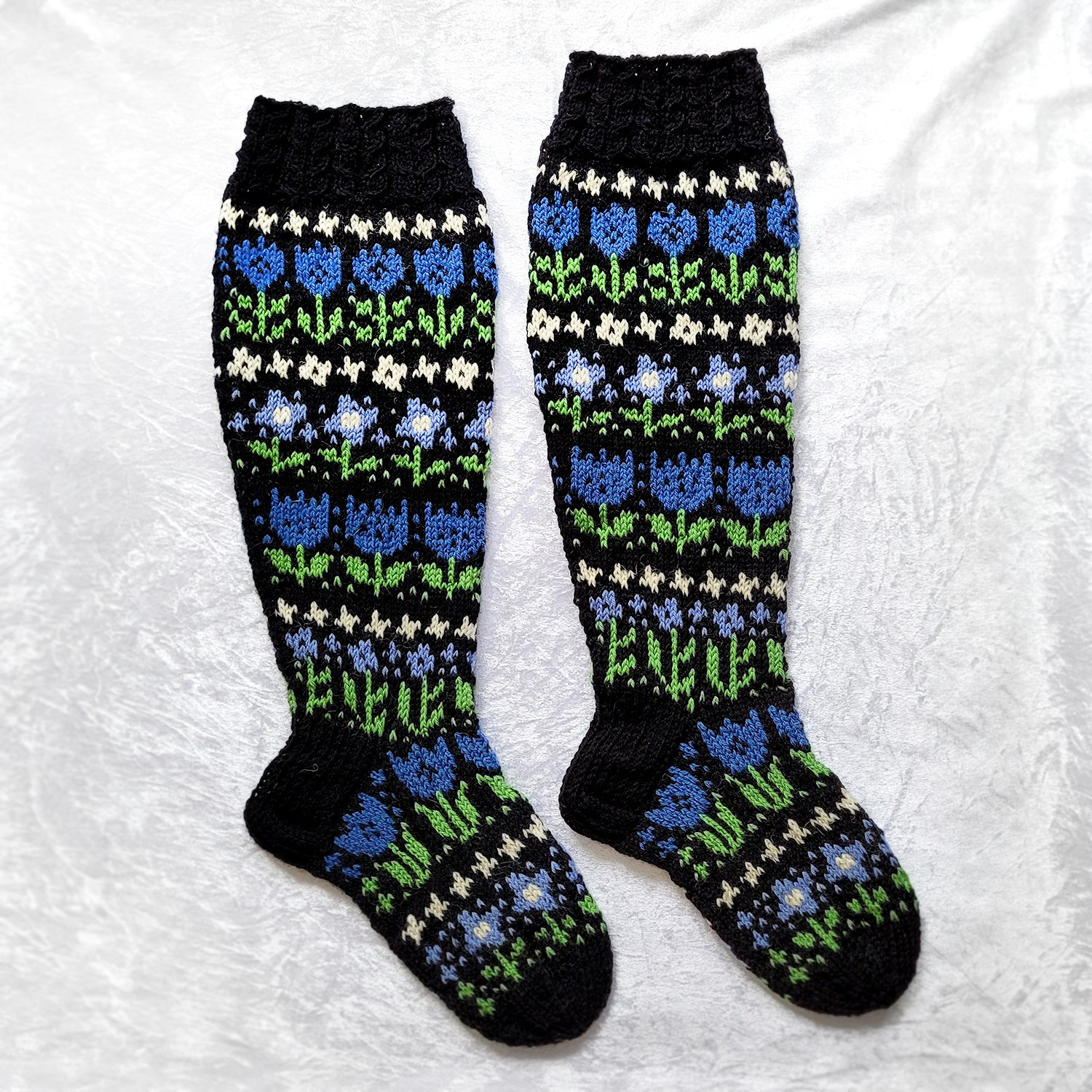 pictured is a pair of knitted knee-high socks, on a light background. they are on a black base, featuring a variety of flowers in two shades of blue, green and white. - 🎨🧶